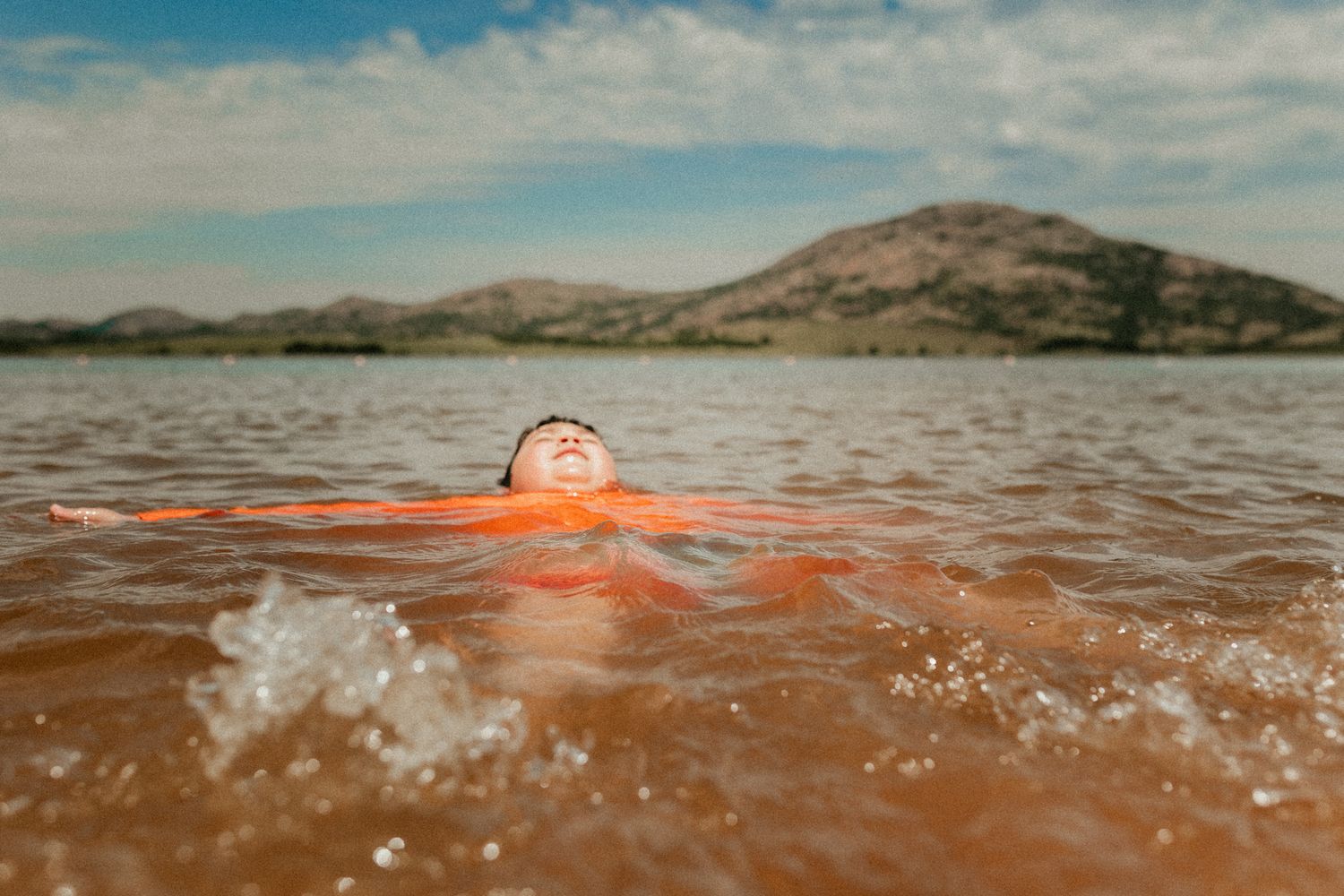 Person floating in brown lake water with mountains in background.