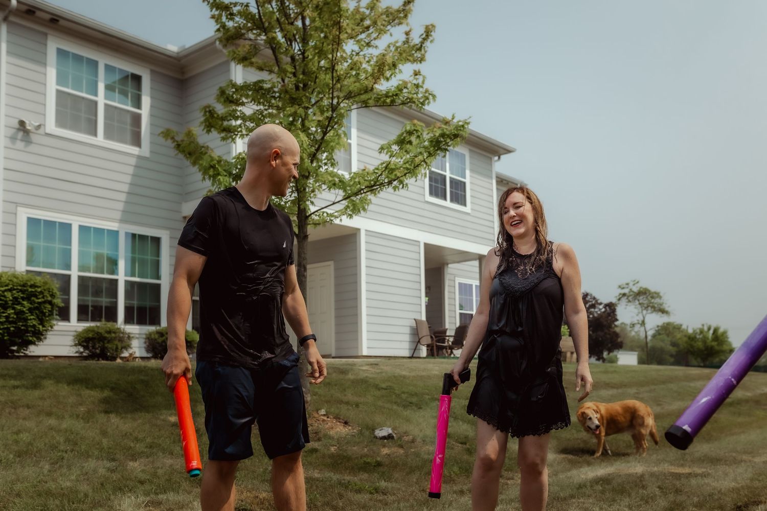 Two people playing with colorful water guns while standing on a lawn with a house in background.