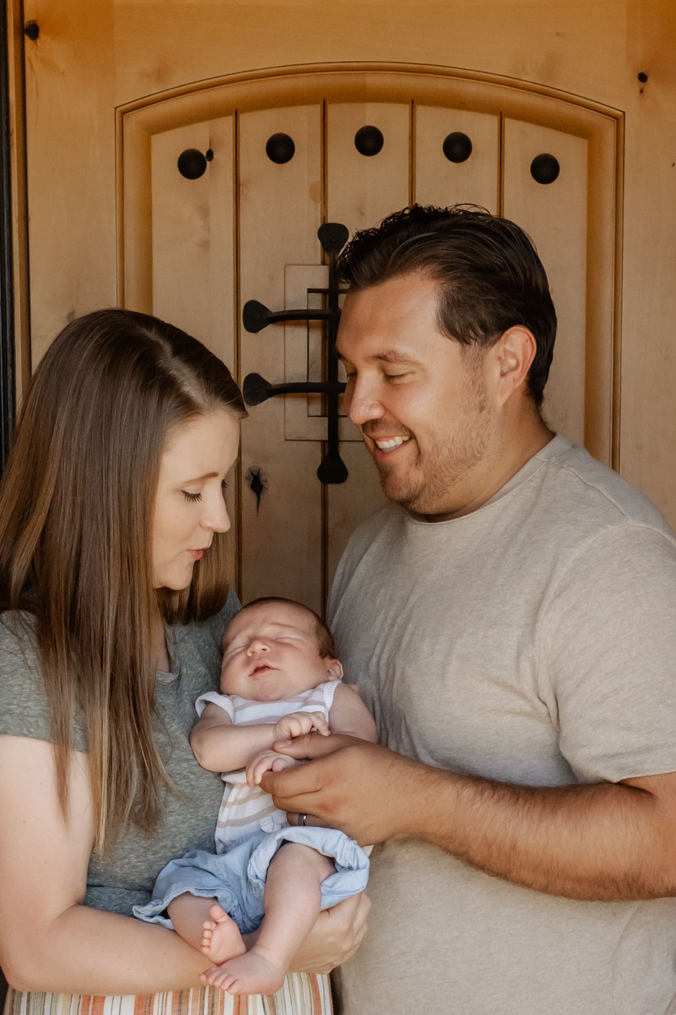 A tender family moment as parents admire their sleeping newborn baby in front of a rustic wooden door.