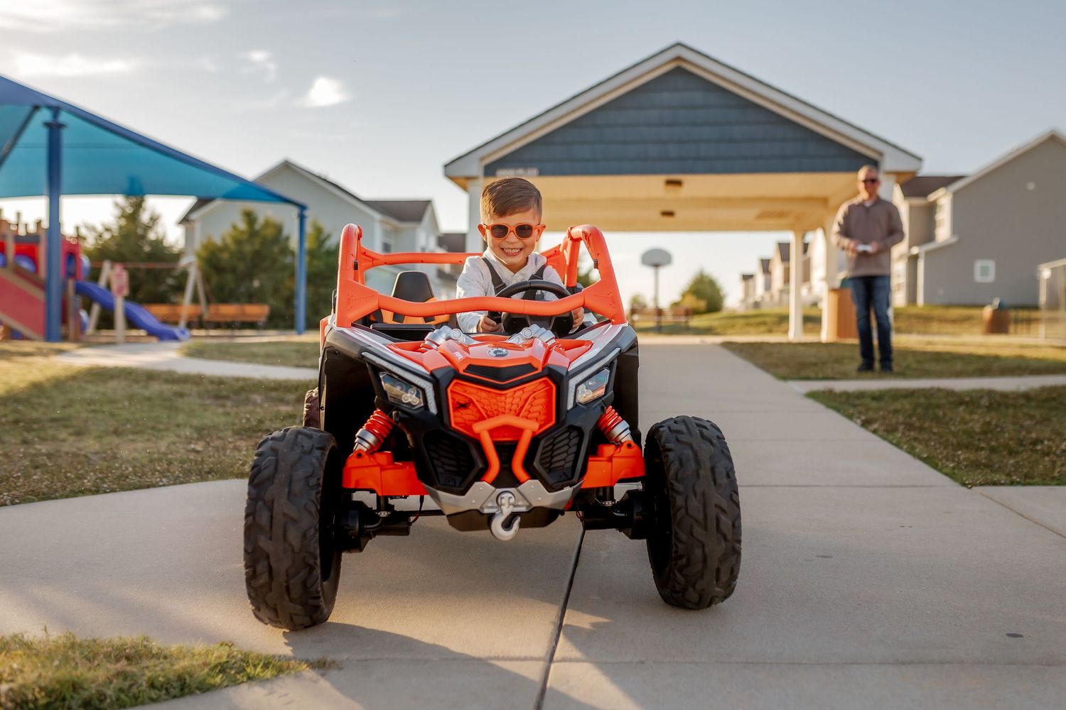 Child driving an orange ATV on a residential sidewalk near playground equipment.
