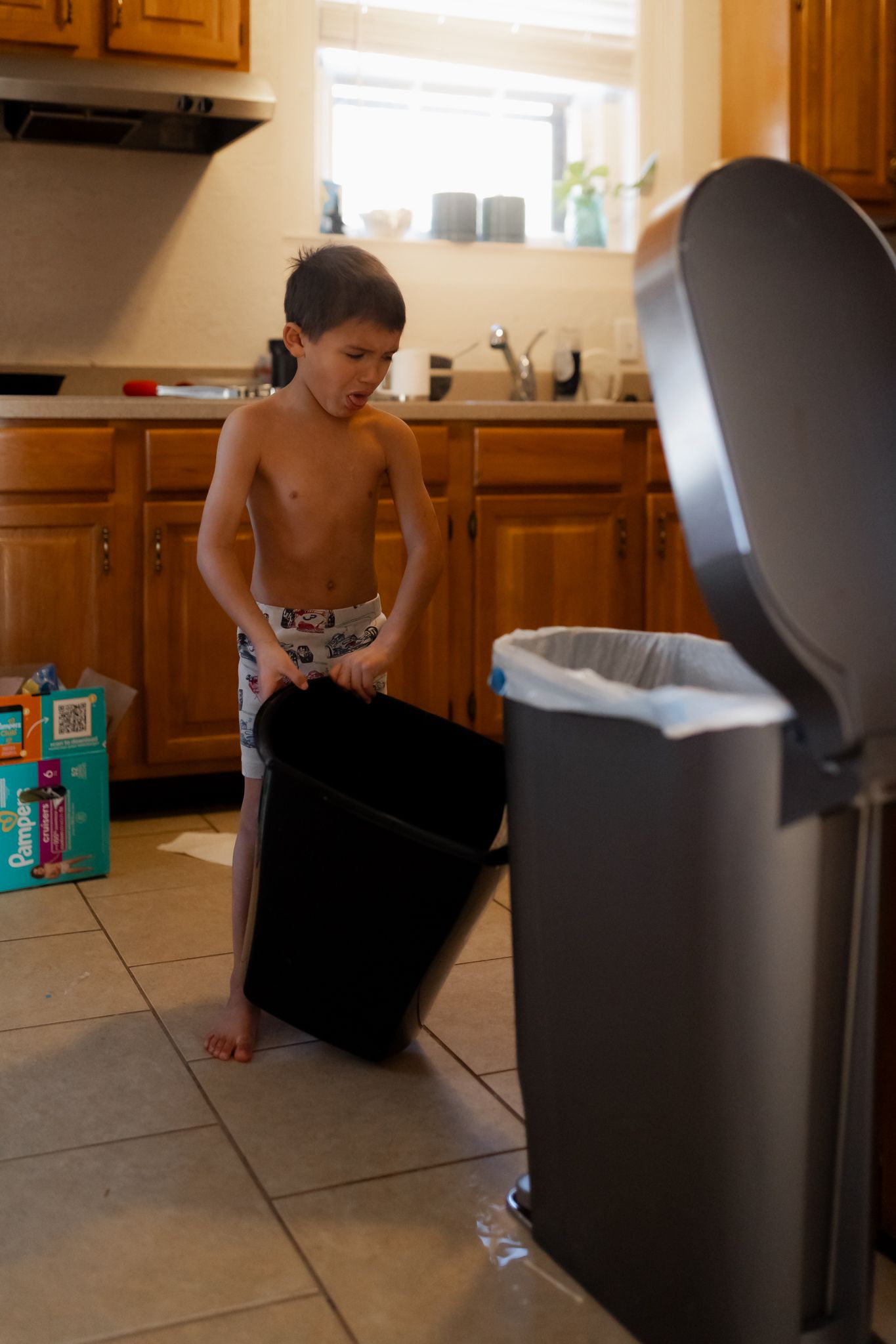 Young child helping with household chores by carrying a large black trash bin in a kitchen.