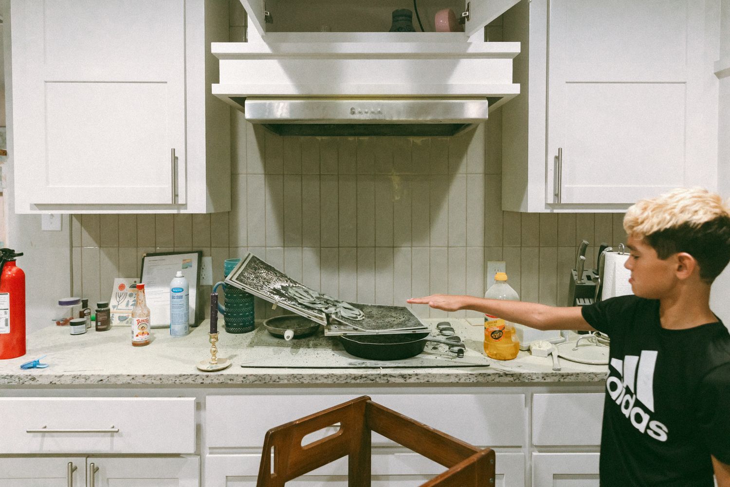Boy in Adidas shirt pointing at stovetop in white kitchen with range hood and cooking pans.
