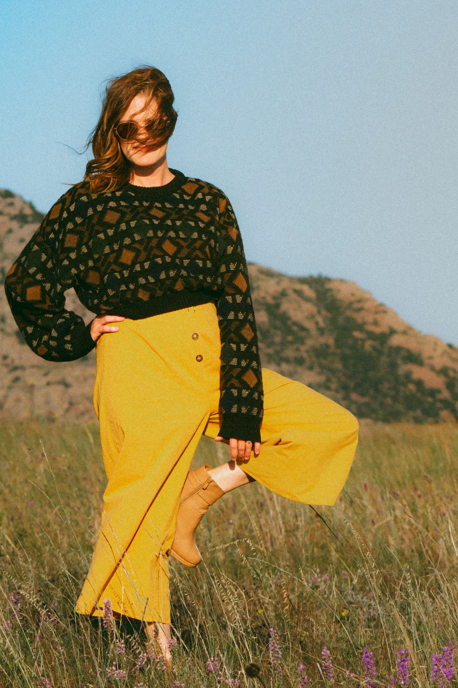 Person wearing black patterned sweater and yellow skirt twirling in mountain meadow with wildflowers.