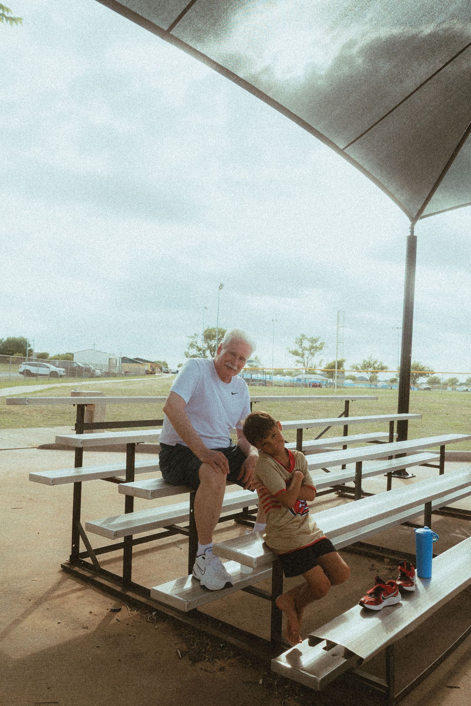 Elderly man and child sitting together on metal bleachers under a covered outdoor sports field shelter.
