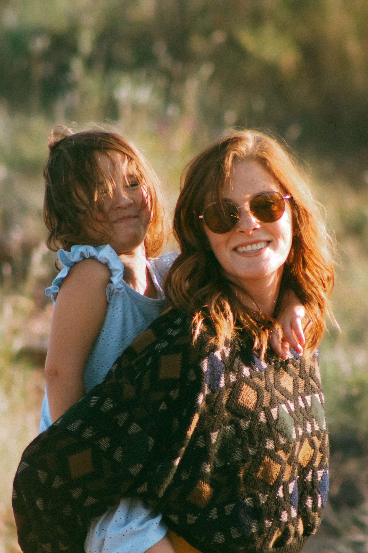 A smiling redhead woman in sunglasses carries a young girl outdoors in warm golden sunlight.