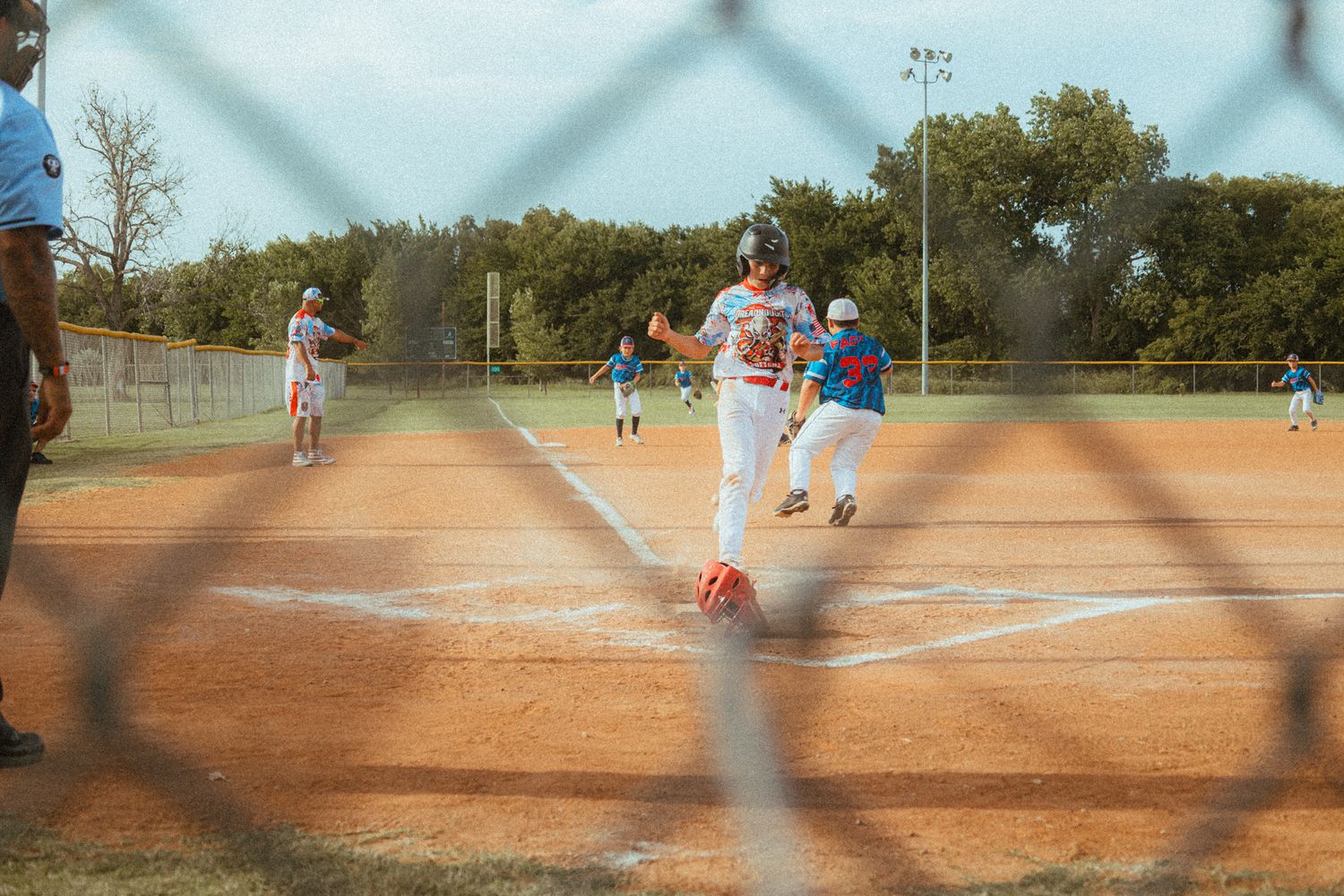 Youth baseball players near home plate on dirt field viewed through chain-link fence.