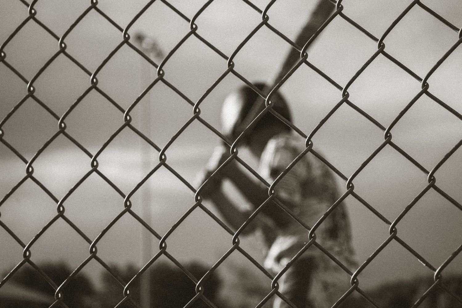 Black and white photo of a baseball player swinging a bat, viewed through a chain-link fence.