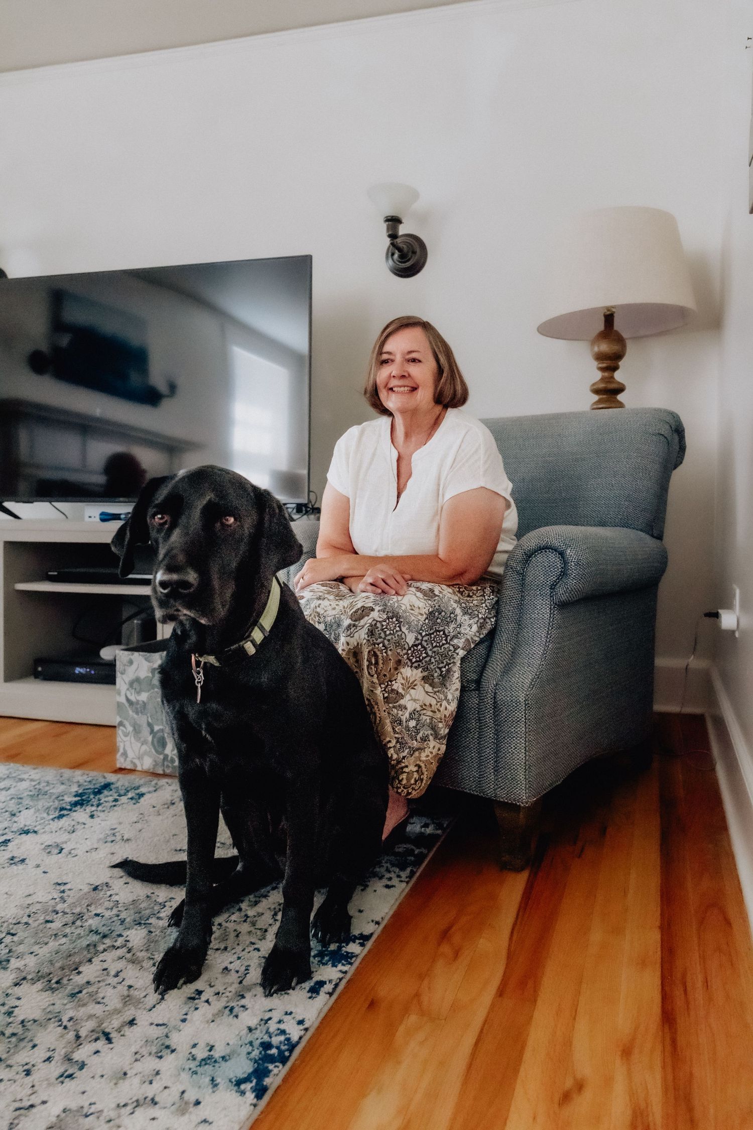Woman smiling in gray armchair beside large black Labrador dog in cozy living room with hardwood floors.