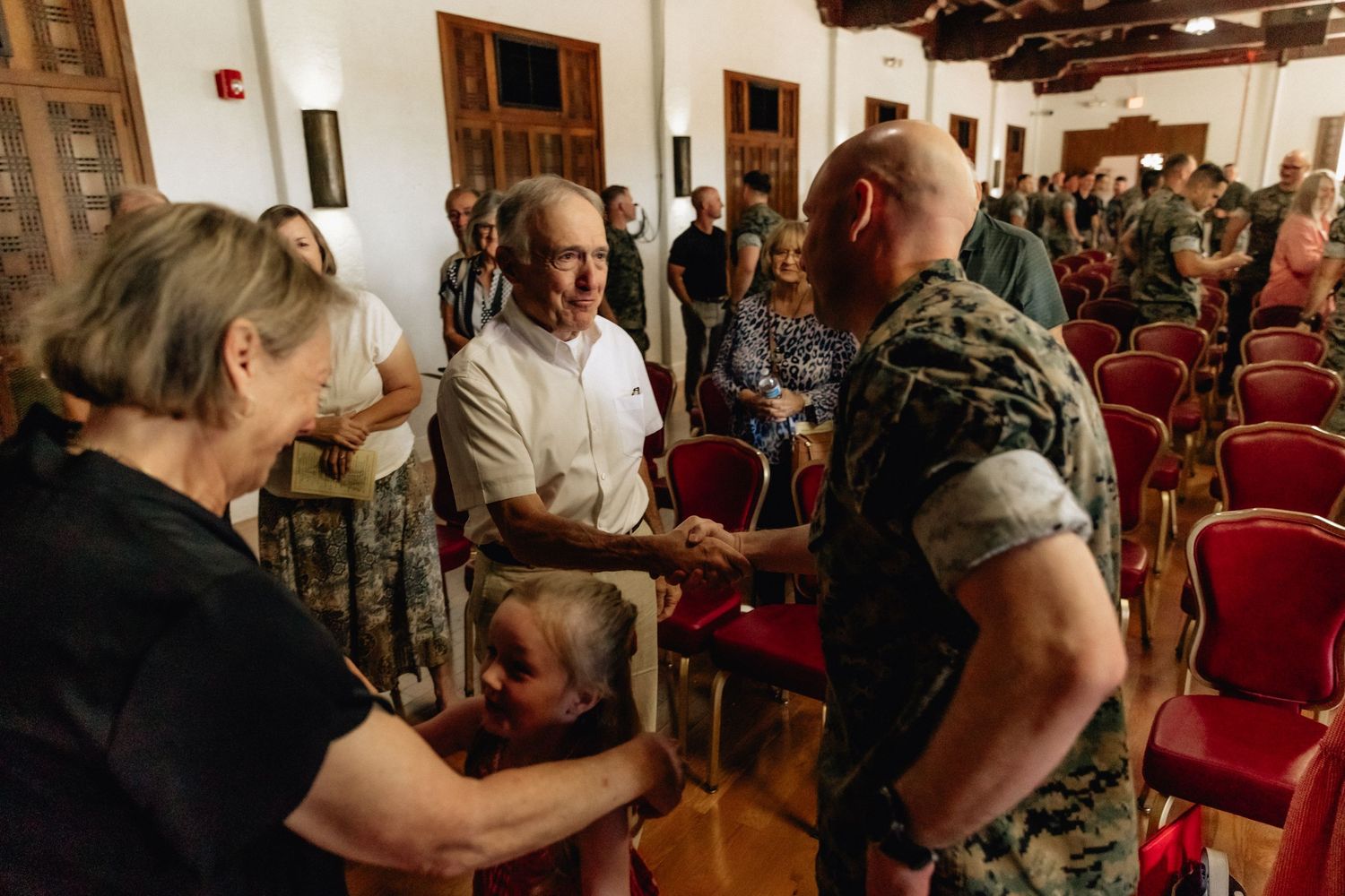 Elderly civilians shake hands with a uniformed Marine soldier inside a hall with red chairs.