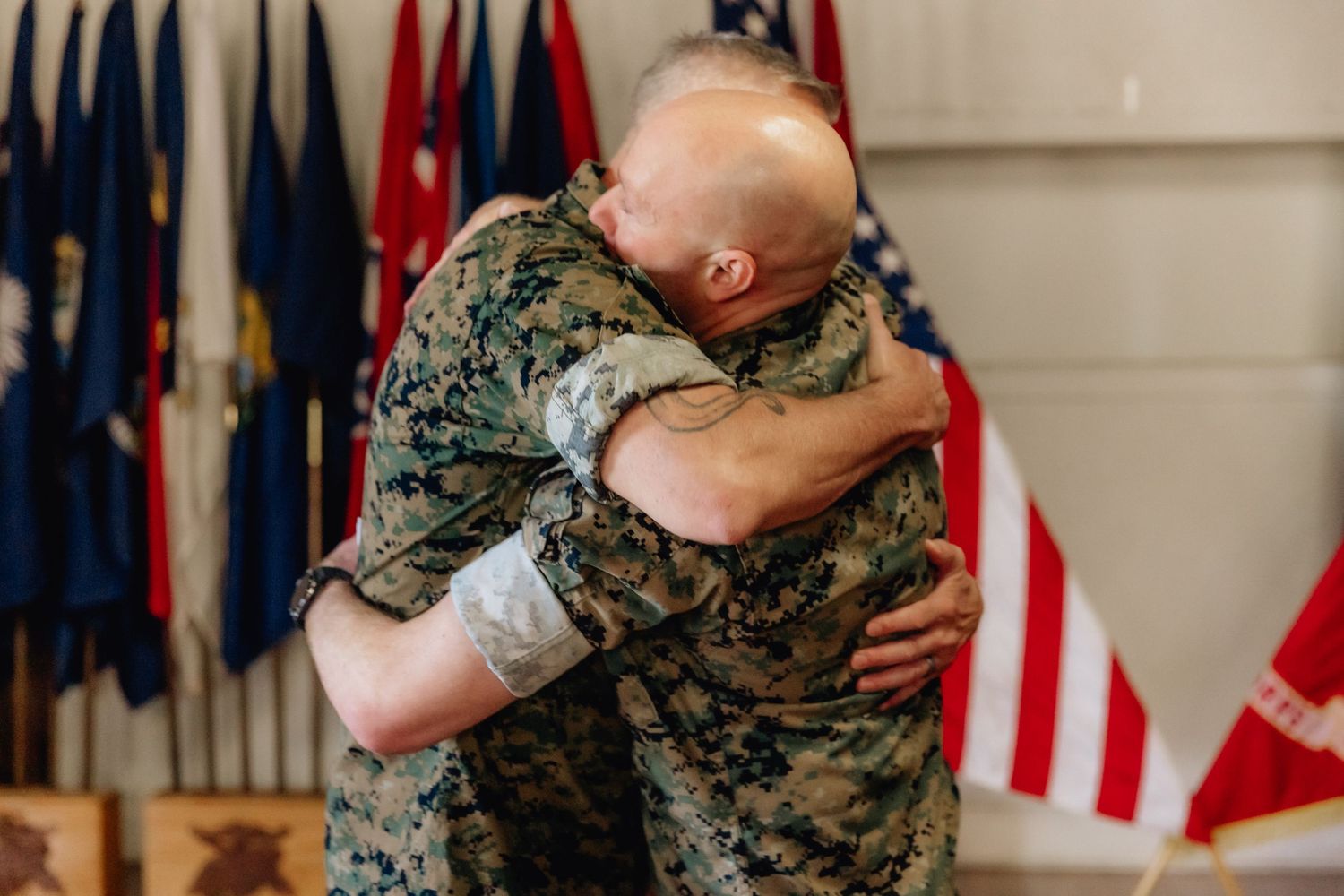 Two Marines in camouflage uniforms share an emotional embrace during a military ceremony with flags in background.