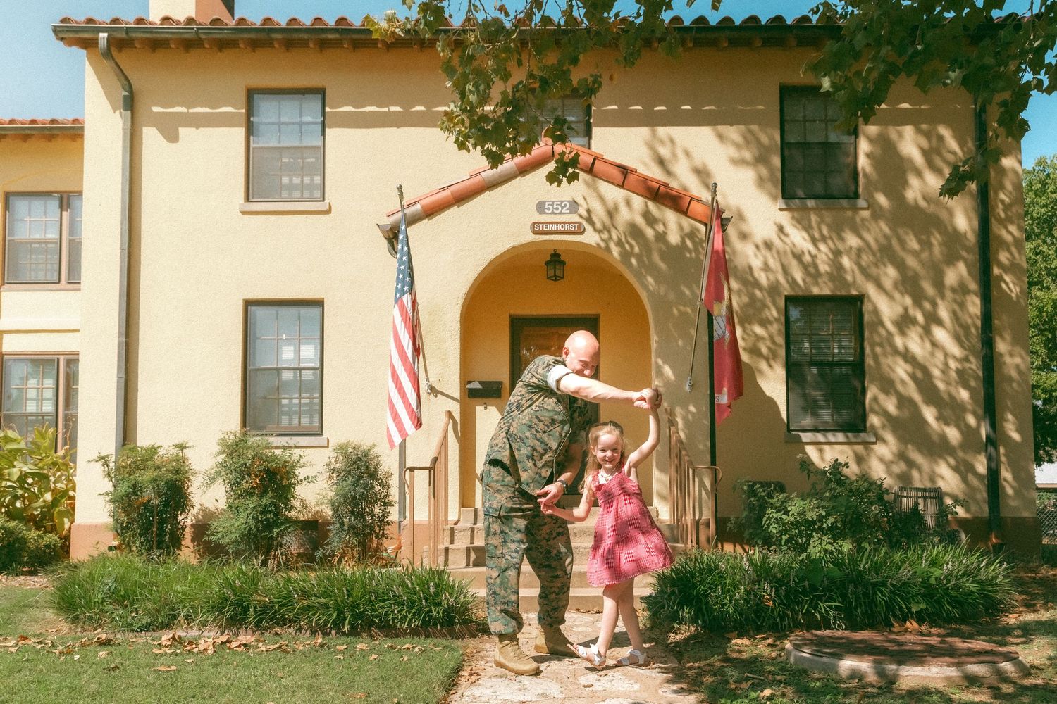 Military soldier reuniting with young daughter outside family home with American flag displayed.