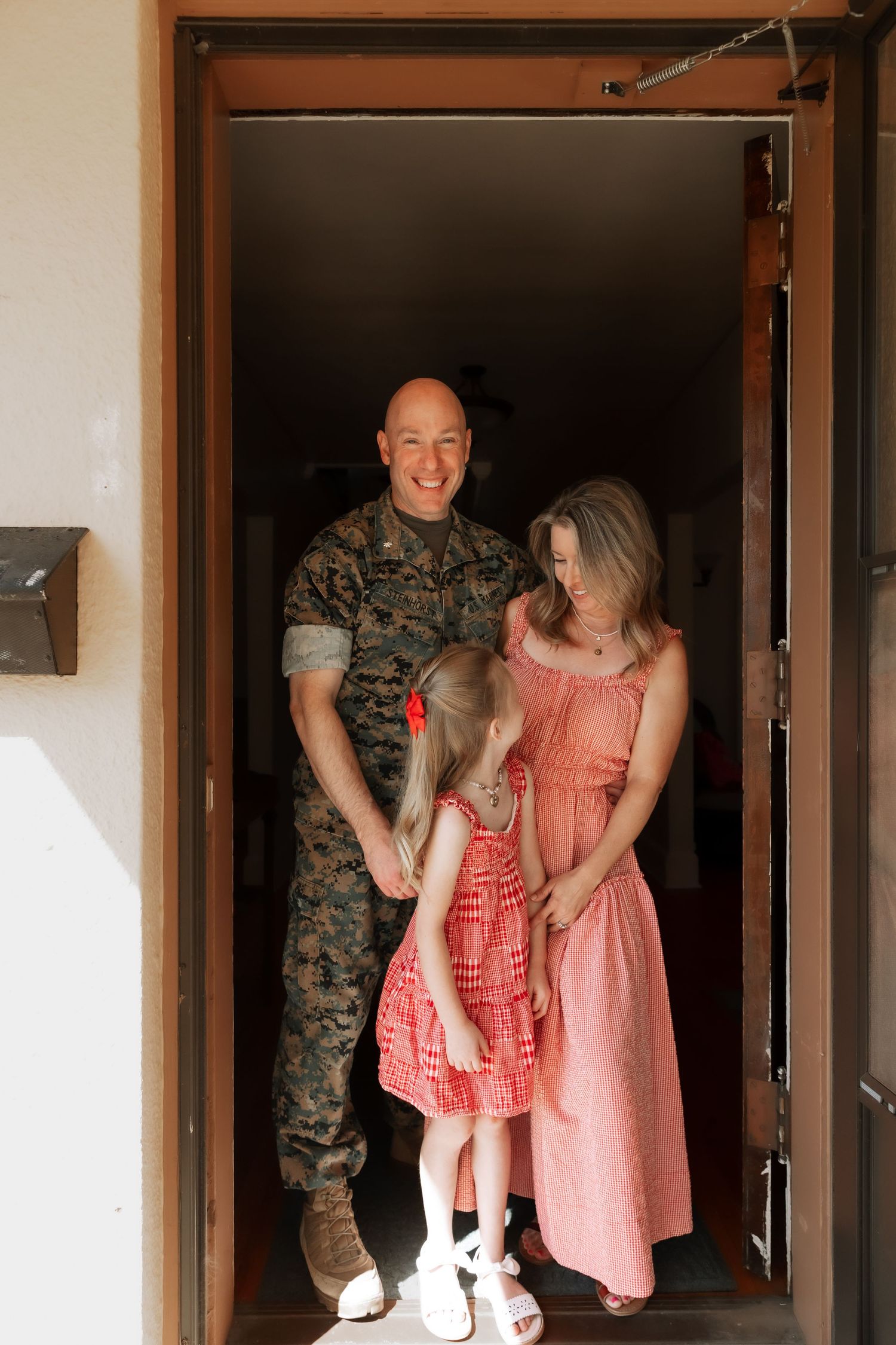 A smiling military man in camouflage uniform stands in a doorway with his wife in a pink dress and young daughter.