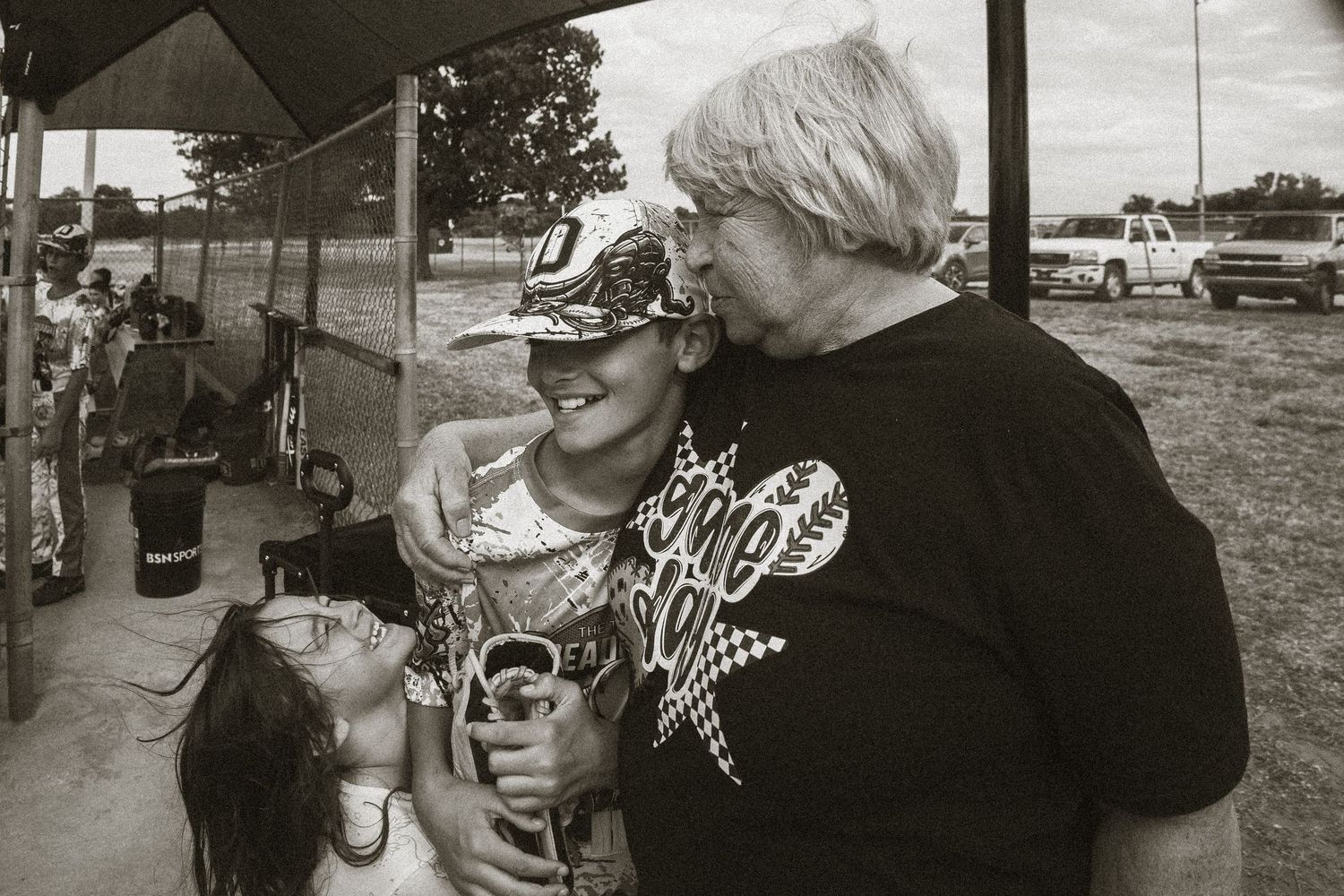 Black and white photo of three people hugging and laughing outdoors at a casual event.