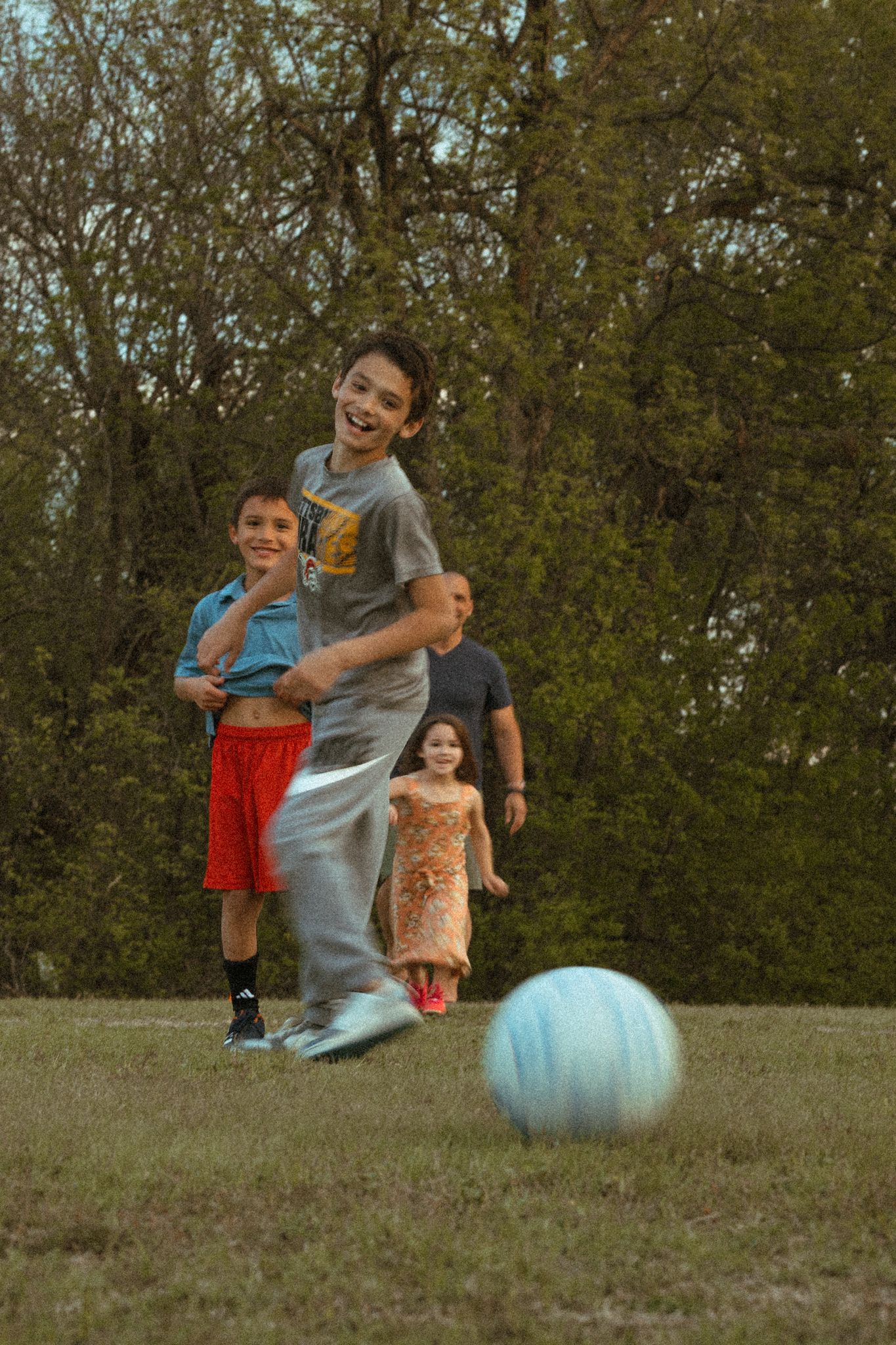 Kids playing soccer in a grassy park, laughing boy kicking blue ball with family watching in background.