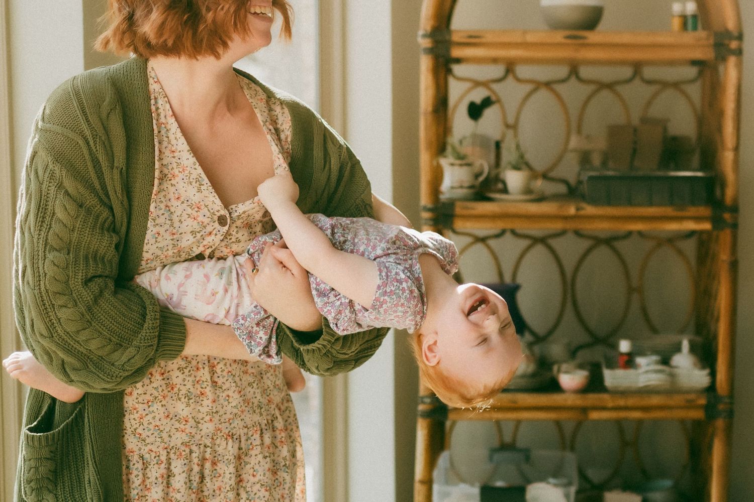 A person in a green cardigan and floral dress stands in a kitchen setting with vintage decor and shelving.