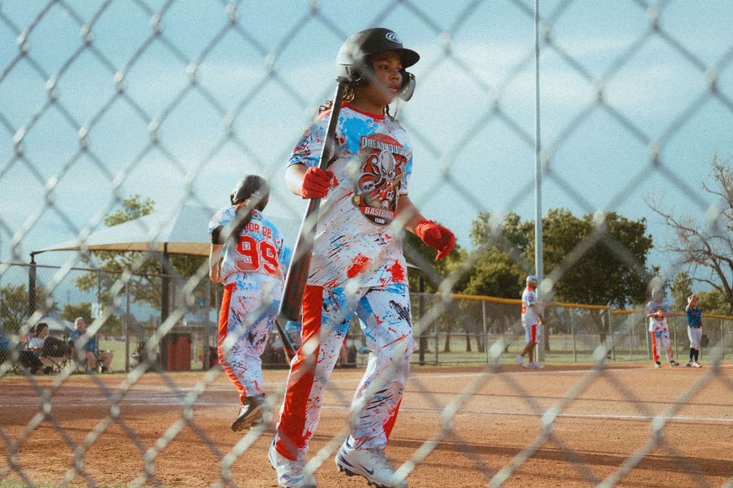 Youth softball player in colorful uniform holding bat behind chain-link fence during game.