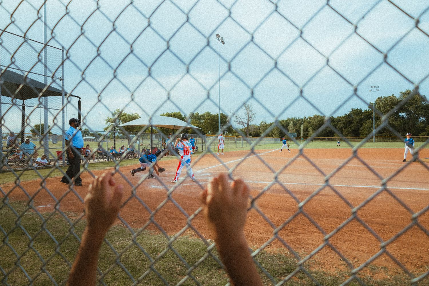 Children playing baseball viewed through a chain-link fence, with players on a dirt diamond field outdoors.