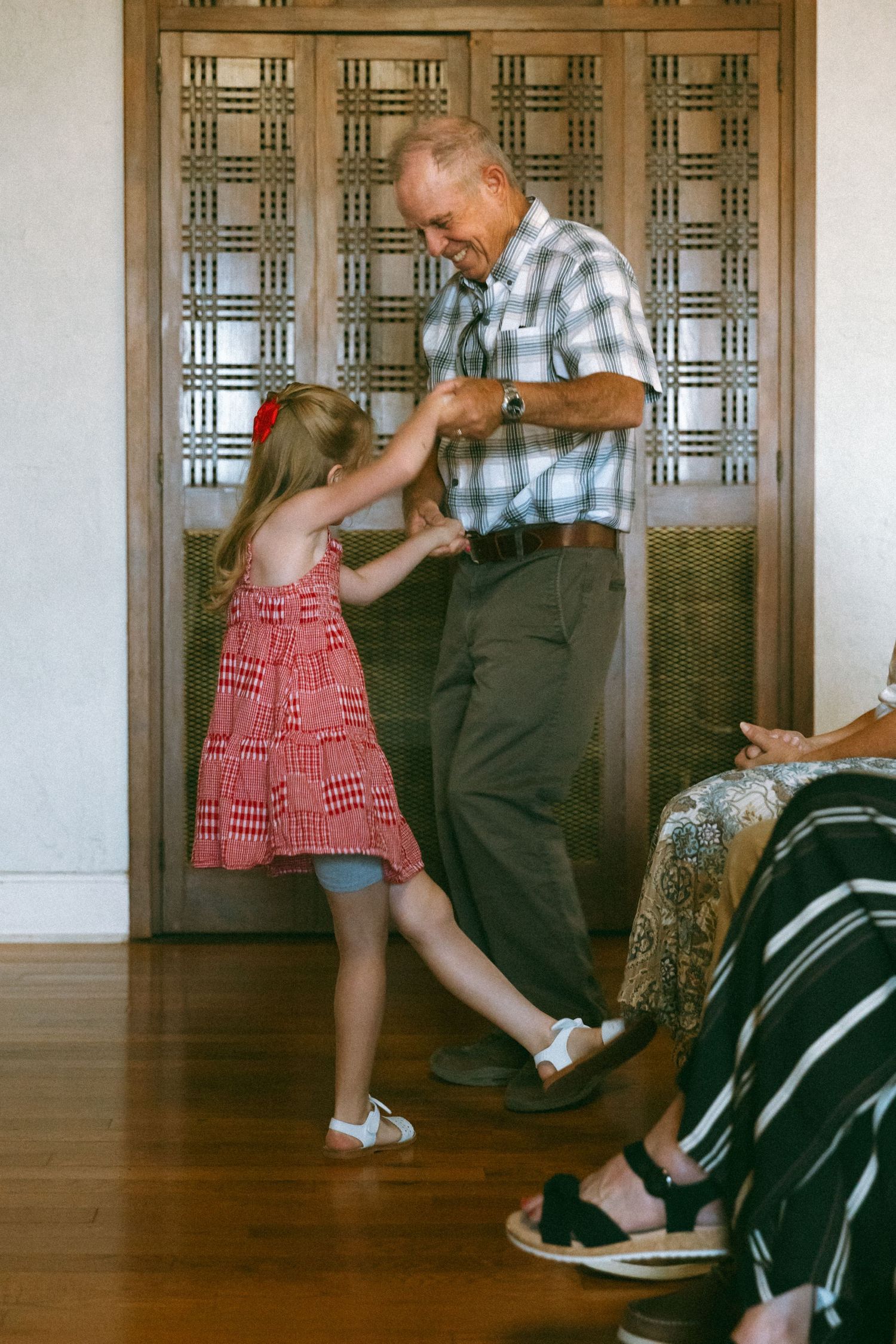 Elderly man dancing with young girl in red dress on hardwood floor indoors.