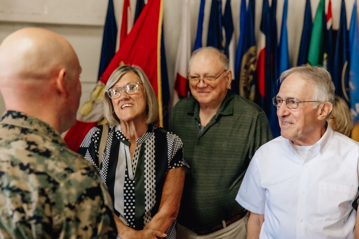 Military officer shaking hands with smiling civilians in front of international flags at a formal indoor event.