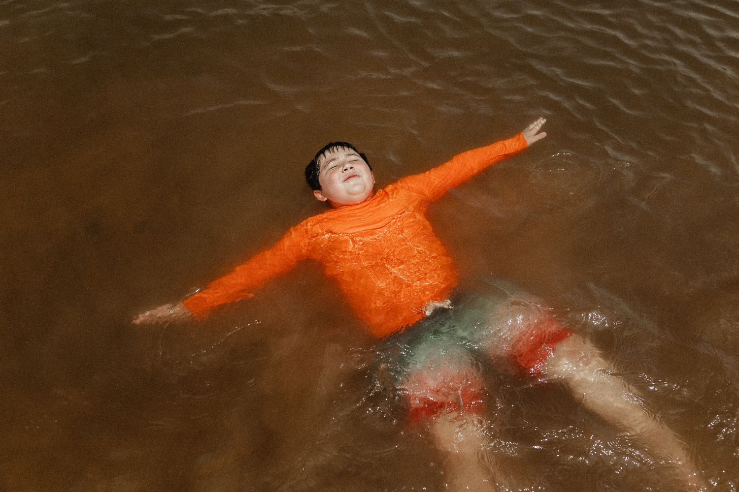 Person in bright orange sweater floating in brown water with arms spread wide.
