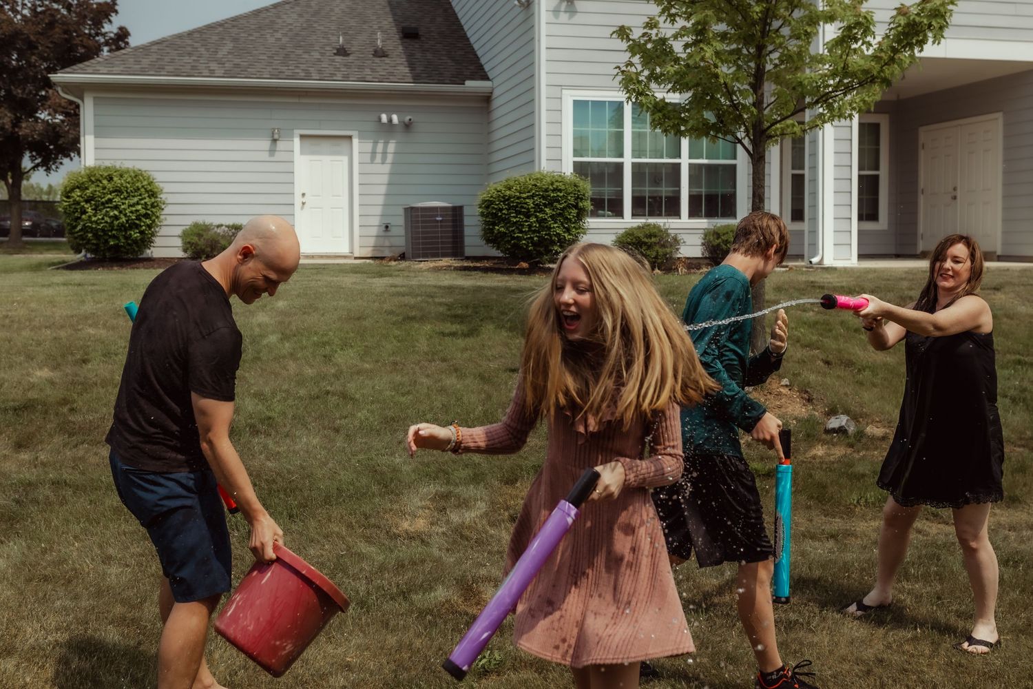 Friends having a playful water fight with buckets and water guns in a residential yard.