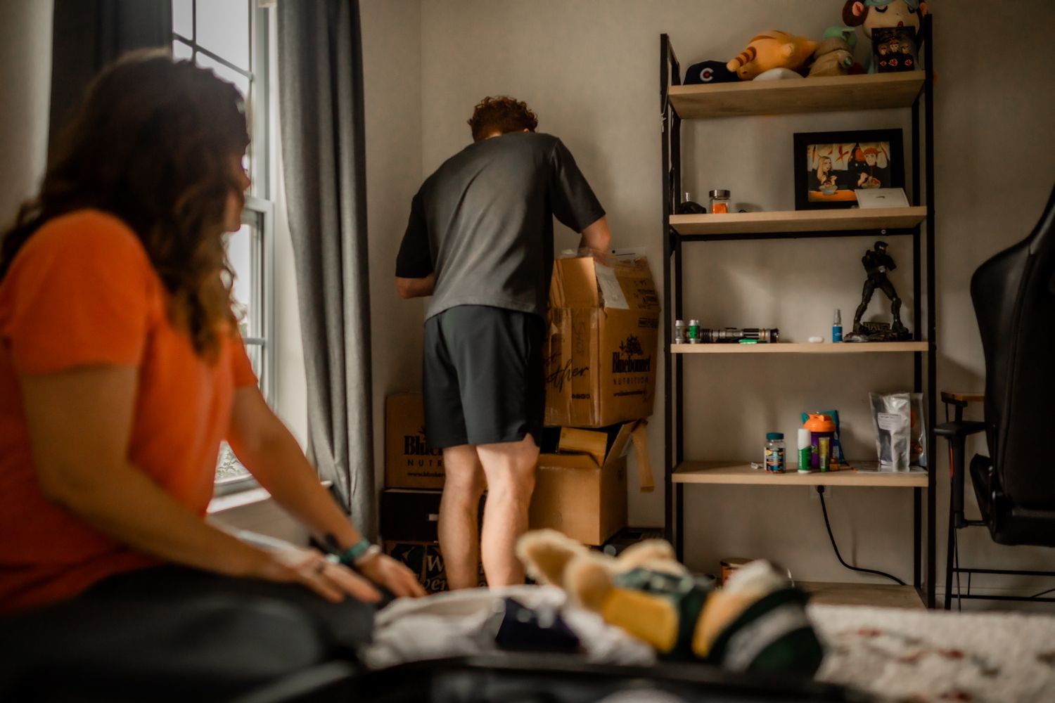 People packing and moving items in bedroom with shelving unit.