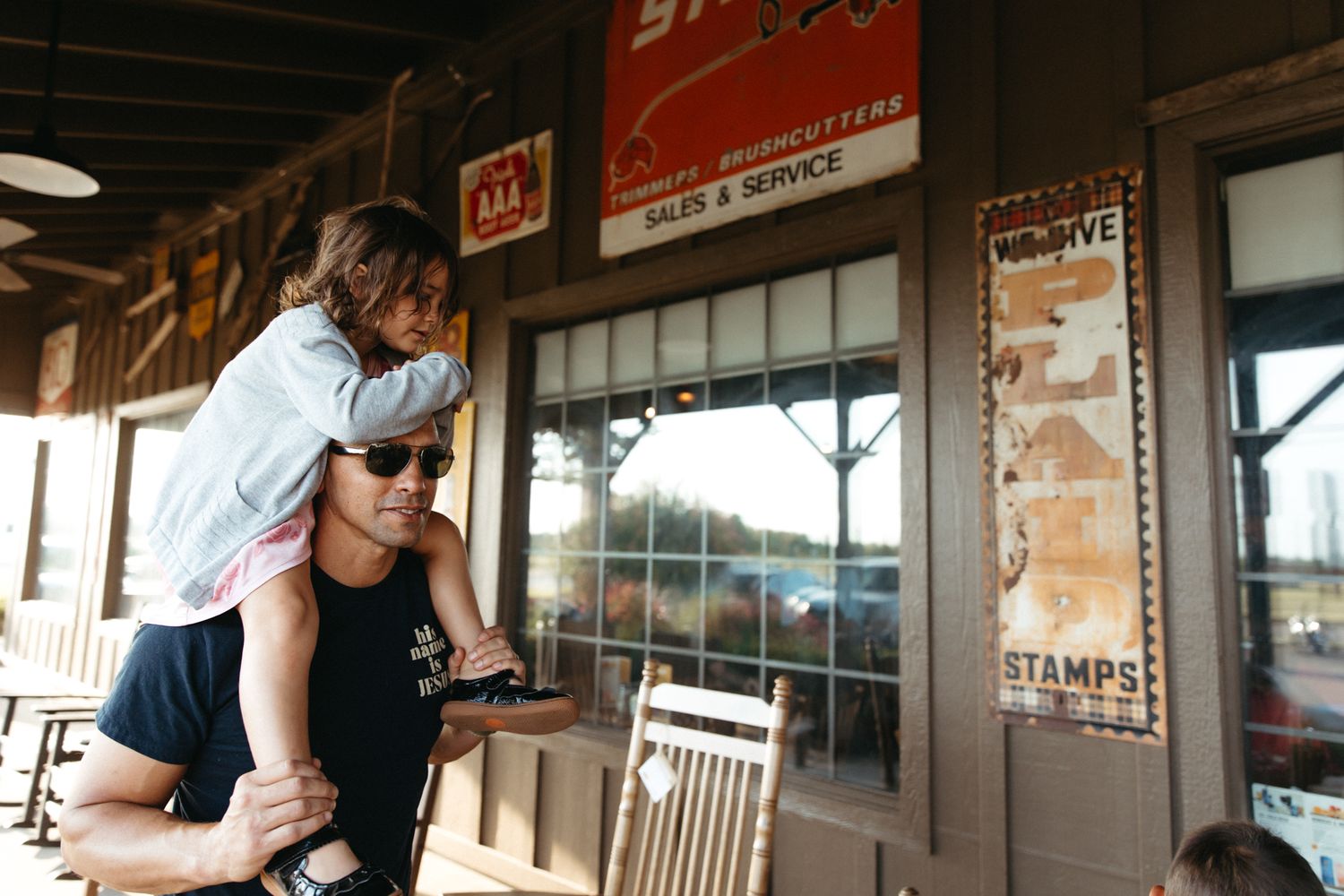 Two people stand together on a covered porch outside a storefront with vintage signs and rocking chairs.