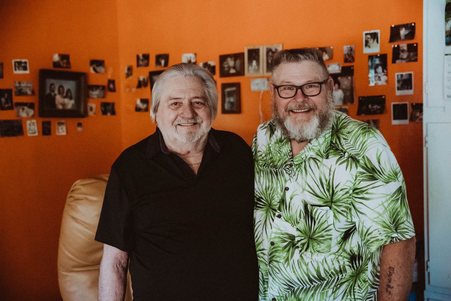 Two senior men standing together against orange wall decorated with photographs.