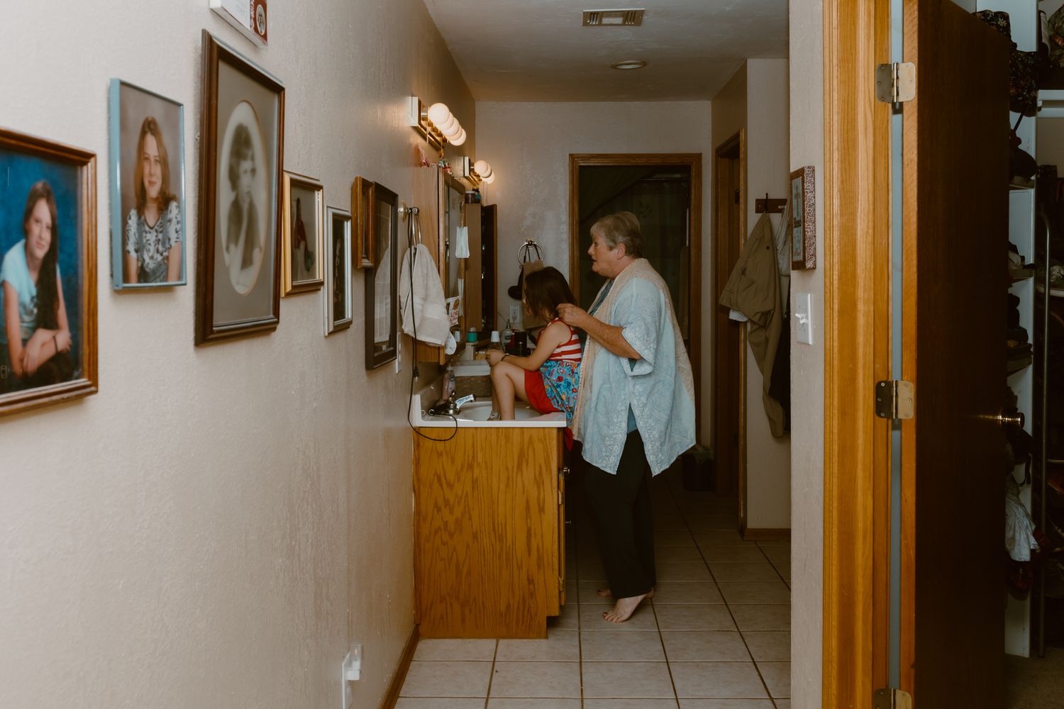 Long hallway with wooden doors and framed photos on beige walls leading to grandma who's fixing a little girls hair in the bathroom mirror.