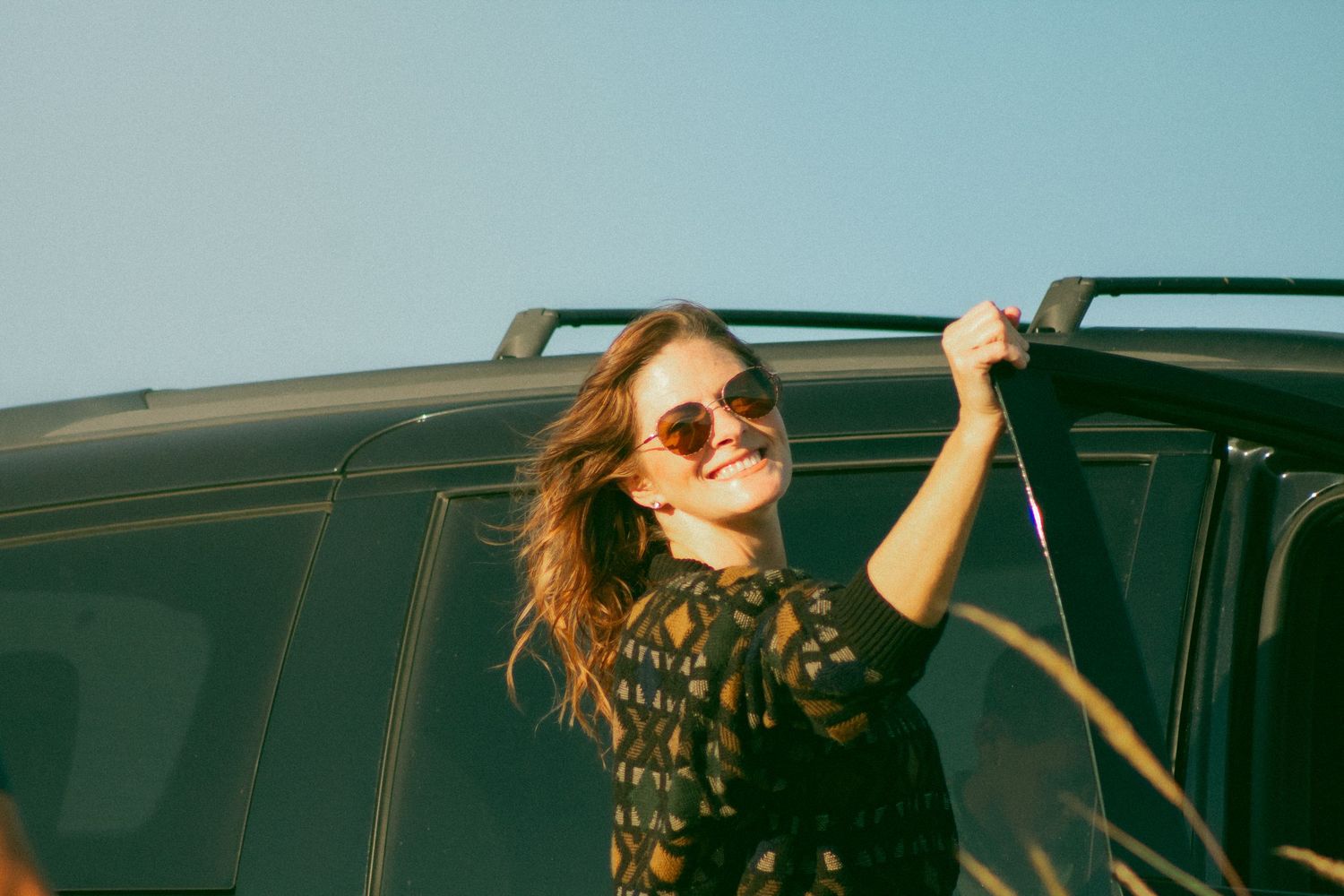 Smiling woman in sunglasses leaning on a dark SUV under a clear blue sky on a sunny day.