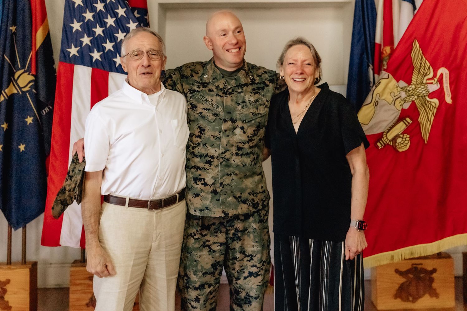 Marine in camouflage uniform poses with two proud family members in front of American and military flags.