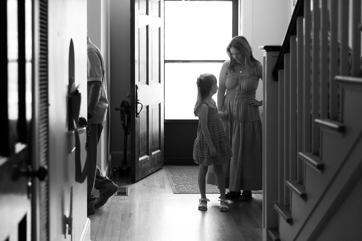 Black and white photo of a mother and young daughter talking in a sunlit home entryway.
