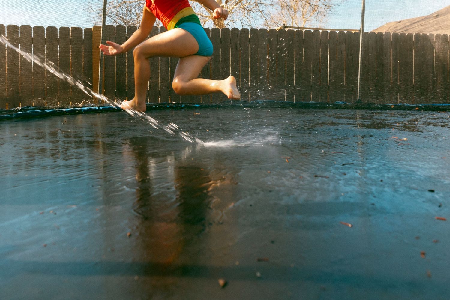 Child jumping on wet trampoline in backyard, water splashing around bare feet on sunny day.