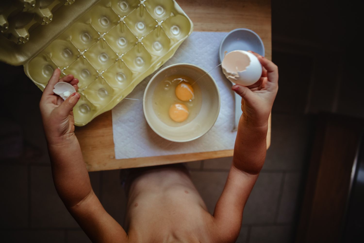 Hands holding egg carton with single egg cracked into white bowl.