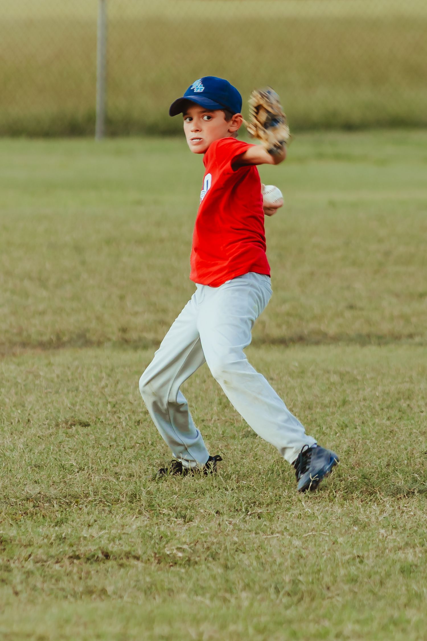 A player in red shirt and baseball cap throwing a game ball across a grassy field during a baseball game. Documentary Photographer Liz Perez