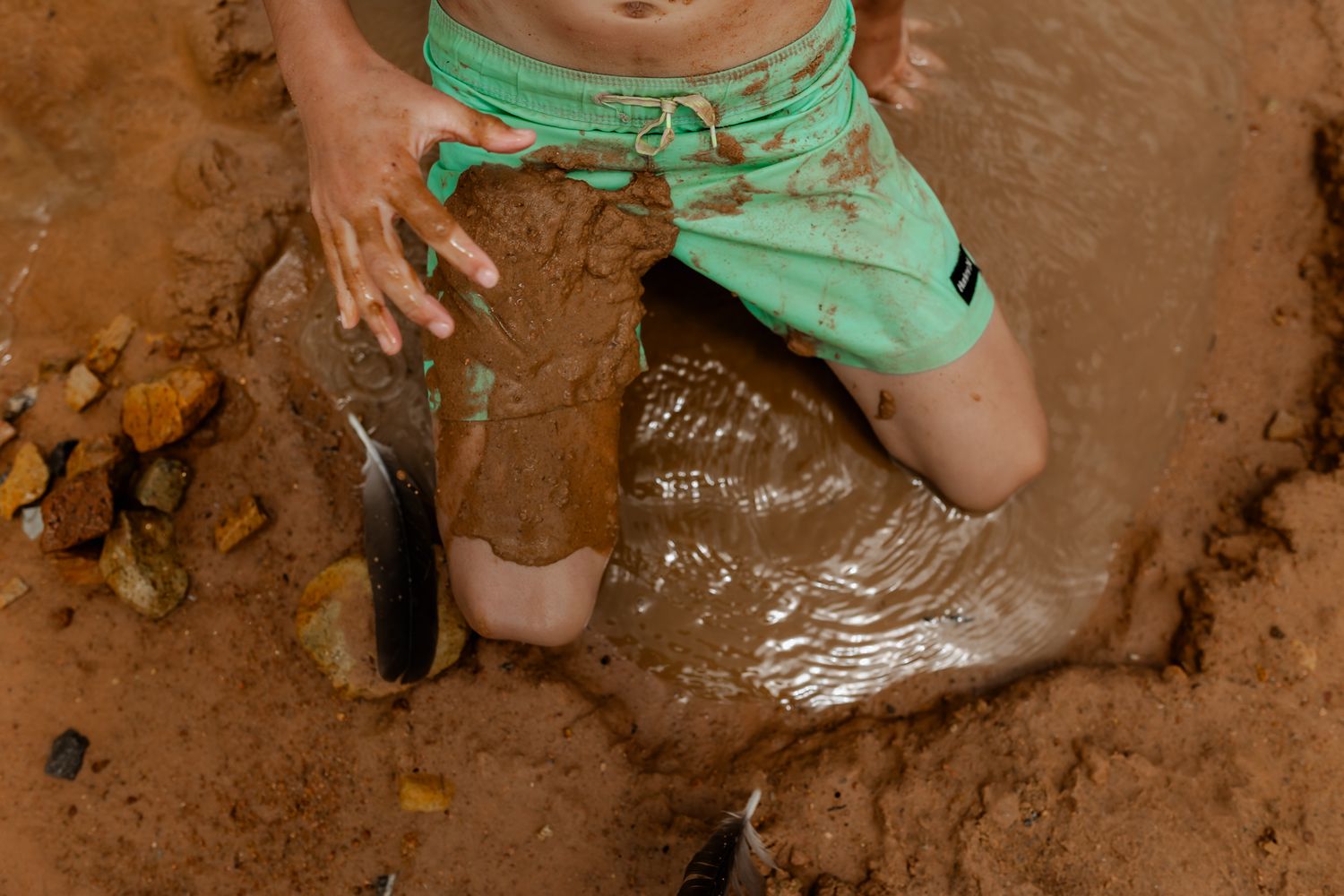 Close-up of legs in mint green shorts sitting in wet muddy sand.