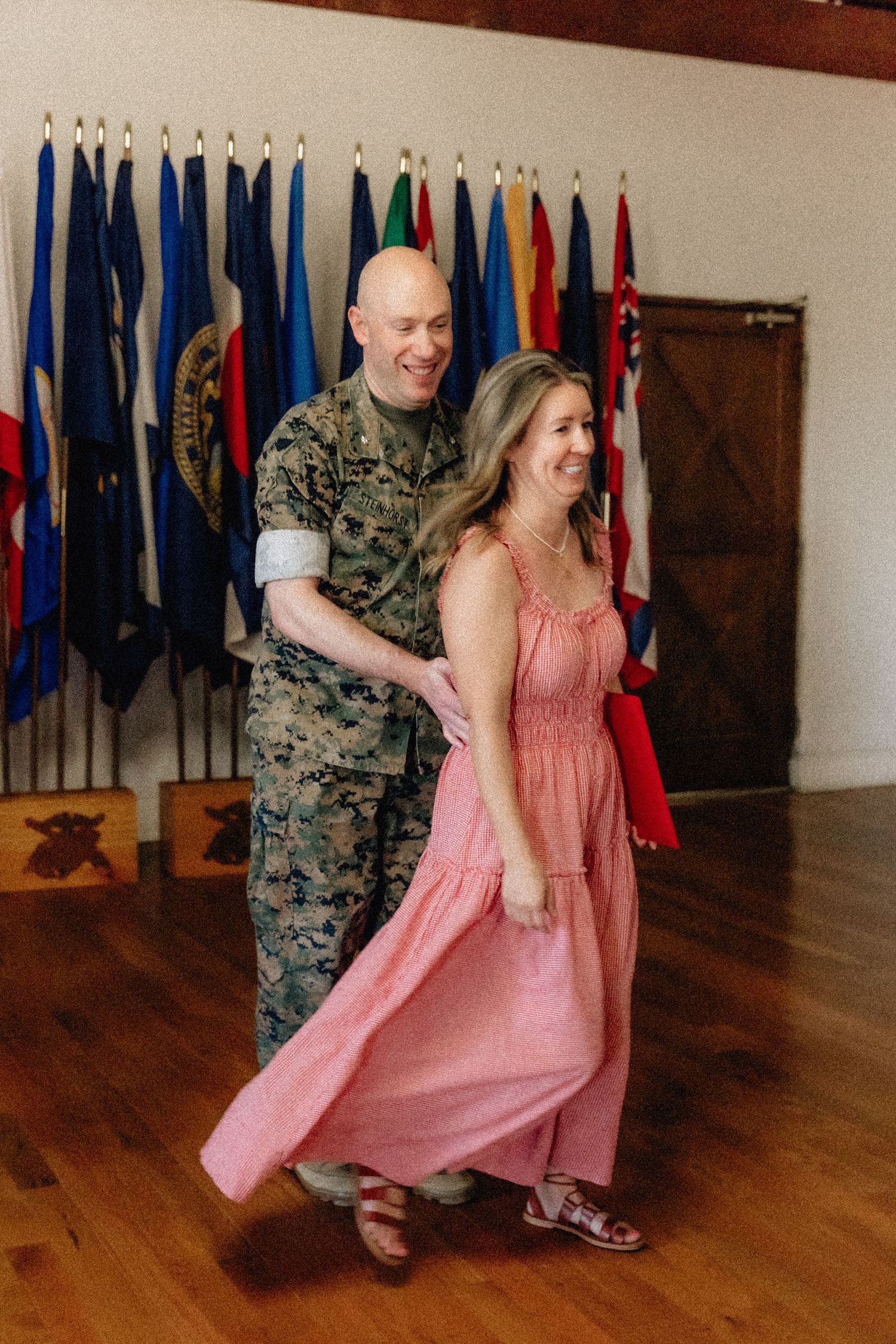 A Marine in uniform dances with a woman in a pink dress before a row of international flags indoors.