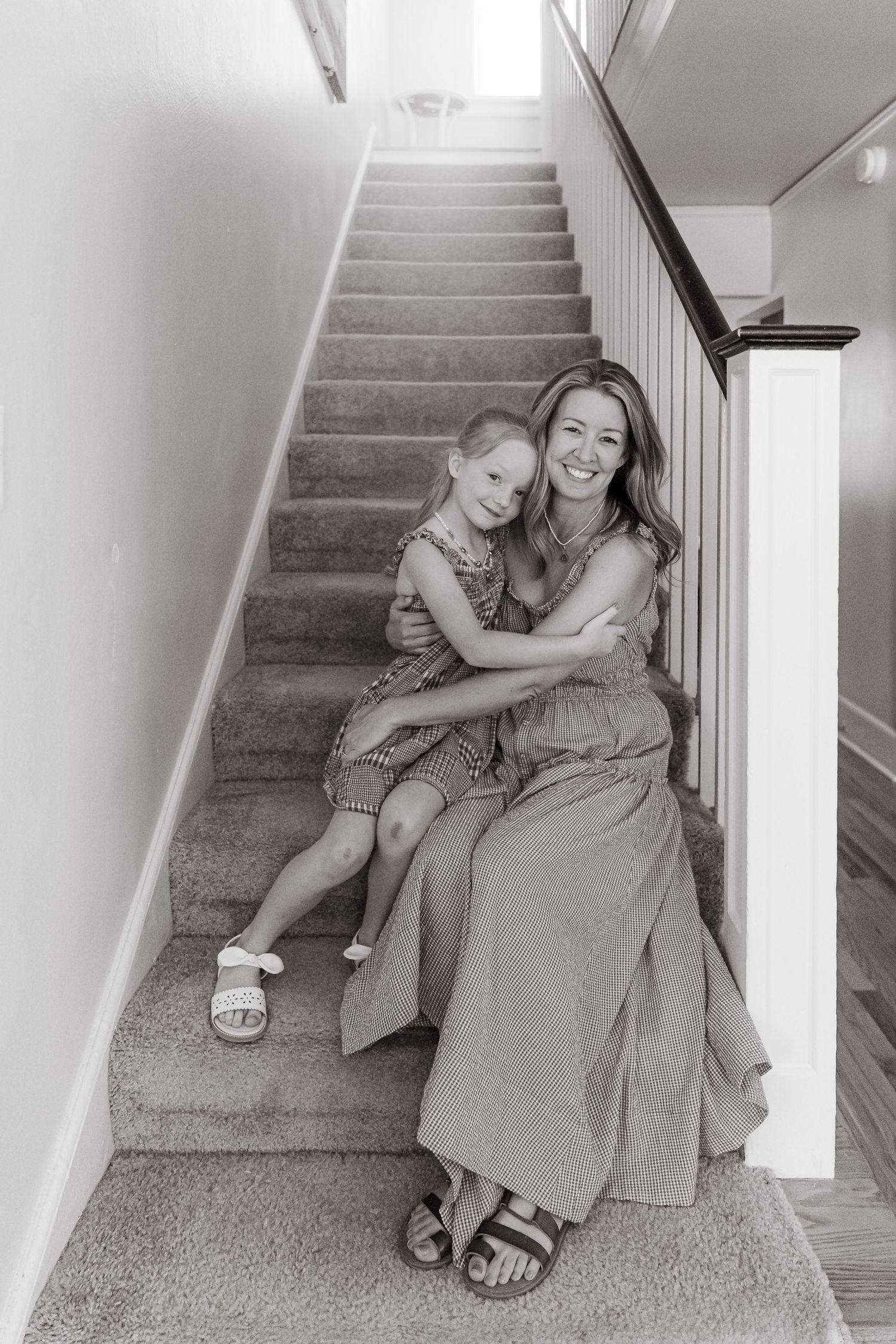Black and white photo of a smiling mother and young daughter hugging on carpeted stairs inside a home.