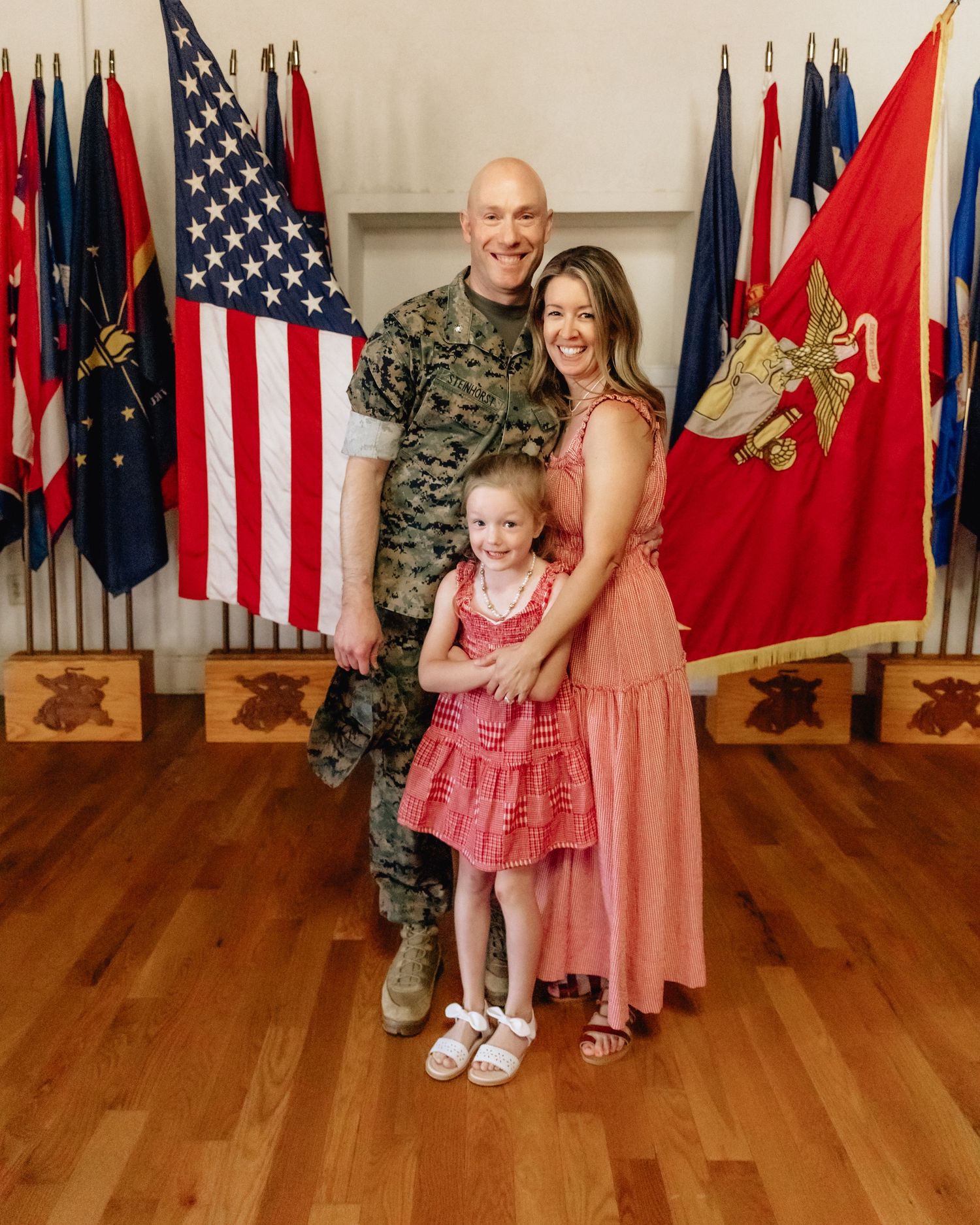 Military family portrait with soldier in camo, wife in pink dress, and young daughter before American flags.
