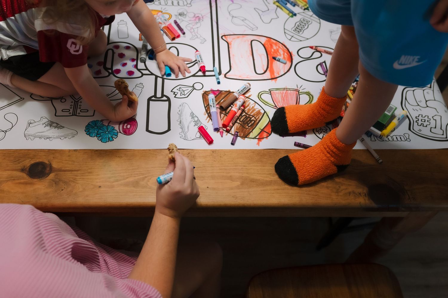 Children coloring a large paper with Crayola markers on a wooden table, viewed from above.