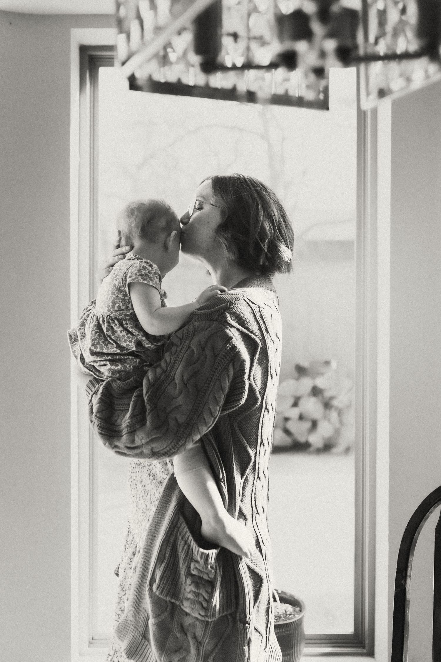 Black and white photograph capturing tender moments by a bright window in a home setting.