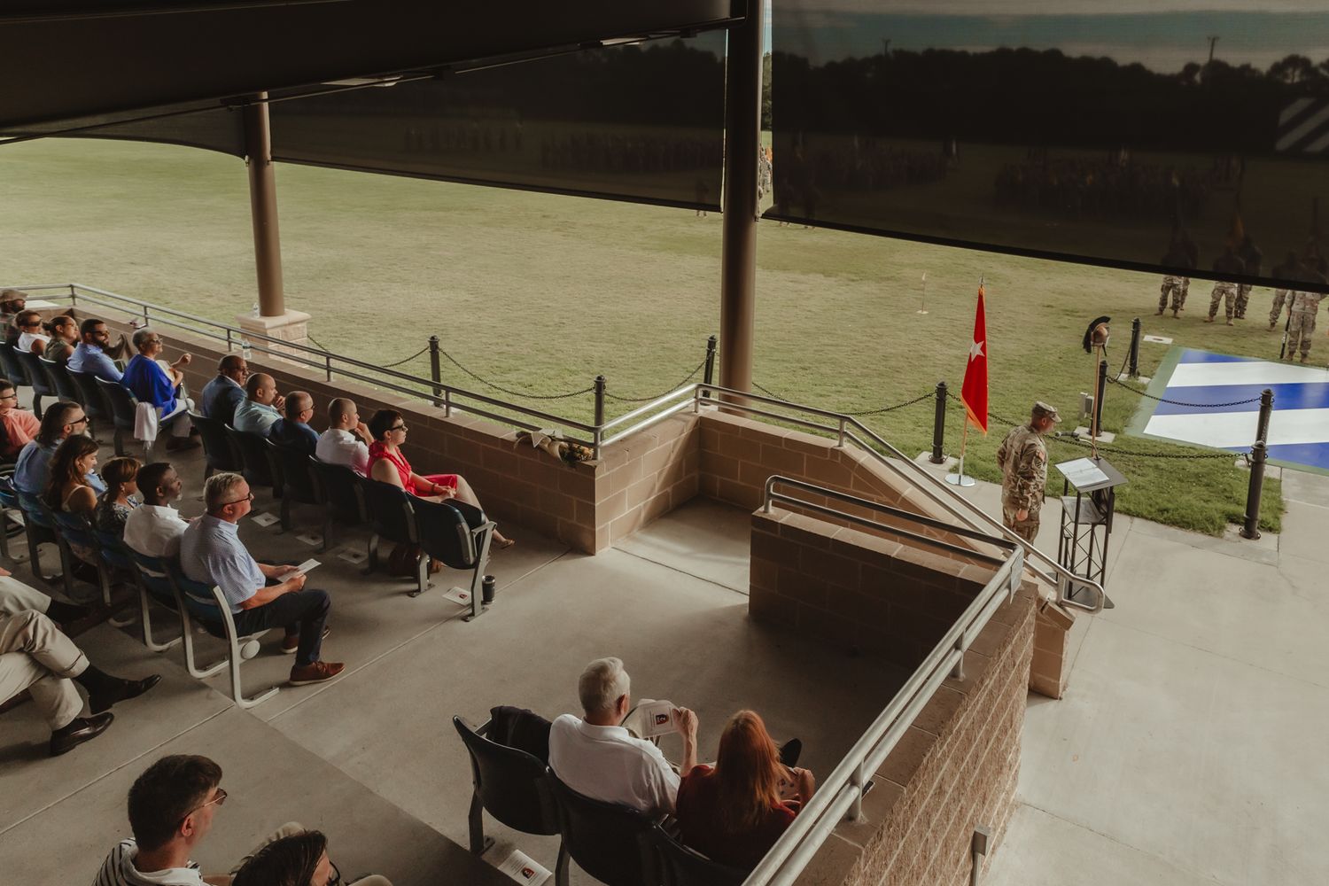 Audience members seated in rows watch a military ceremony from covered bleachers.