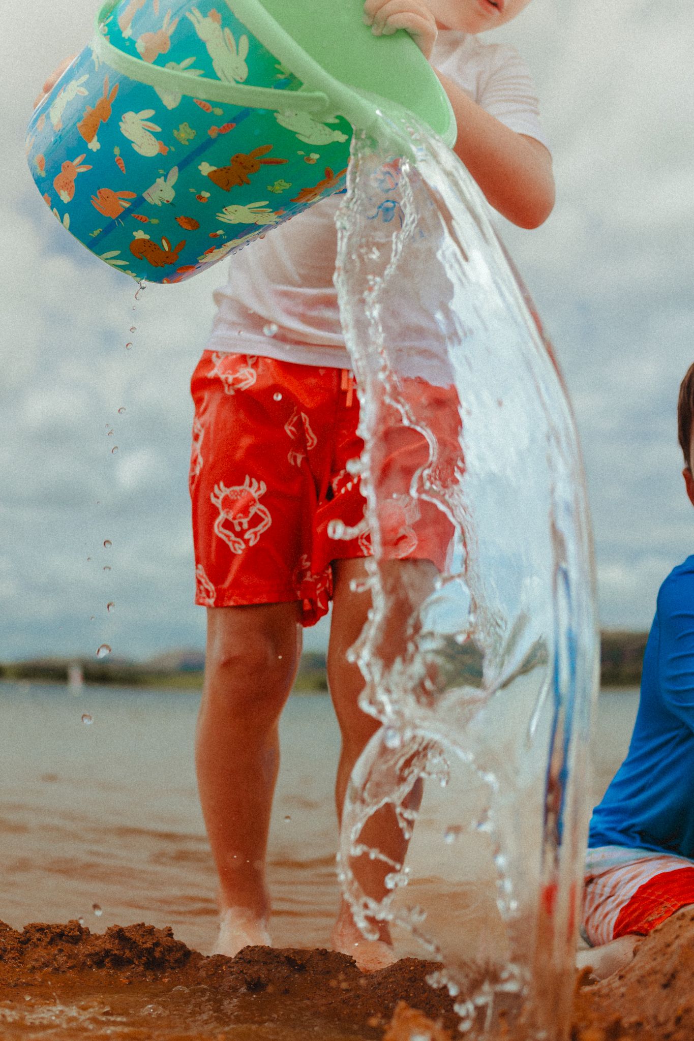 Water pouring from turquoise bucket creating a splash near children playing in sand.