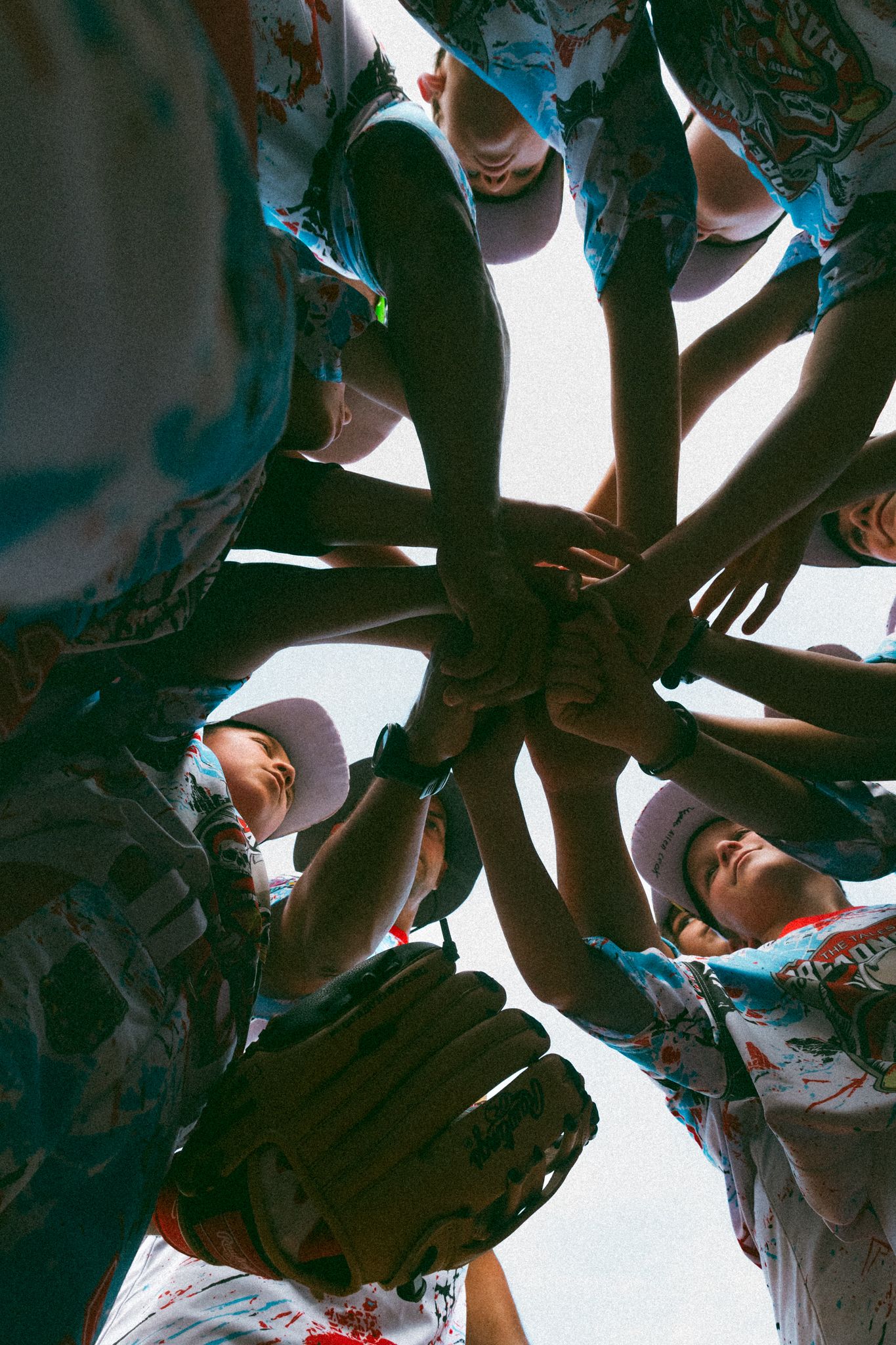 Youth baseball team huddles in a circle stacking hands together, viewed from below, wearing matching uniforms and caps.