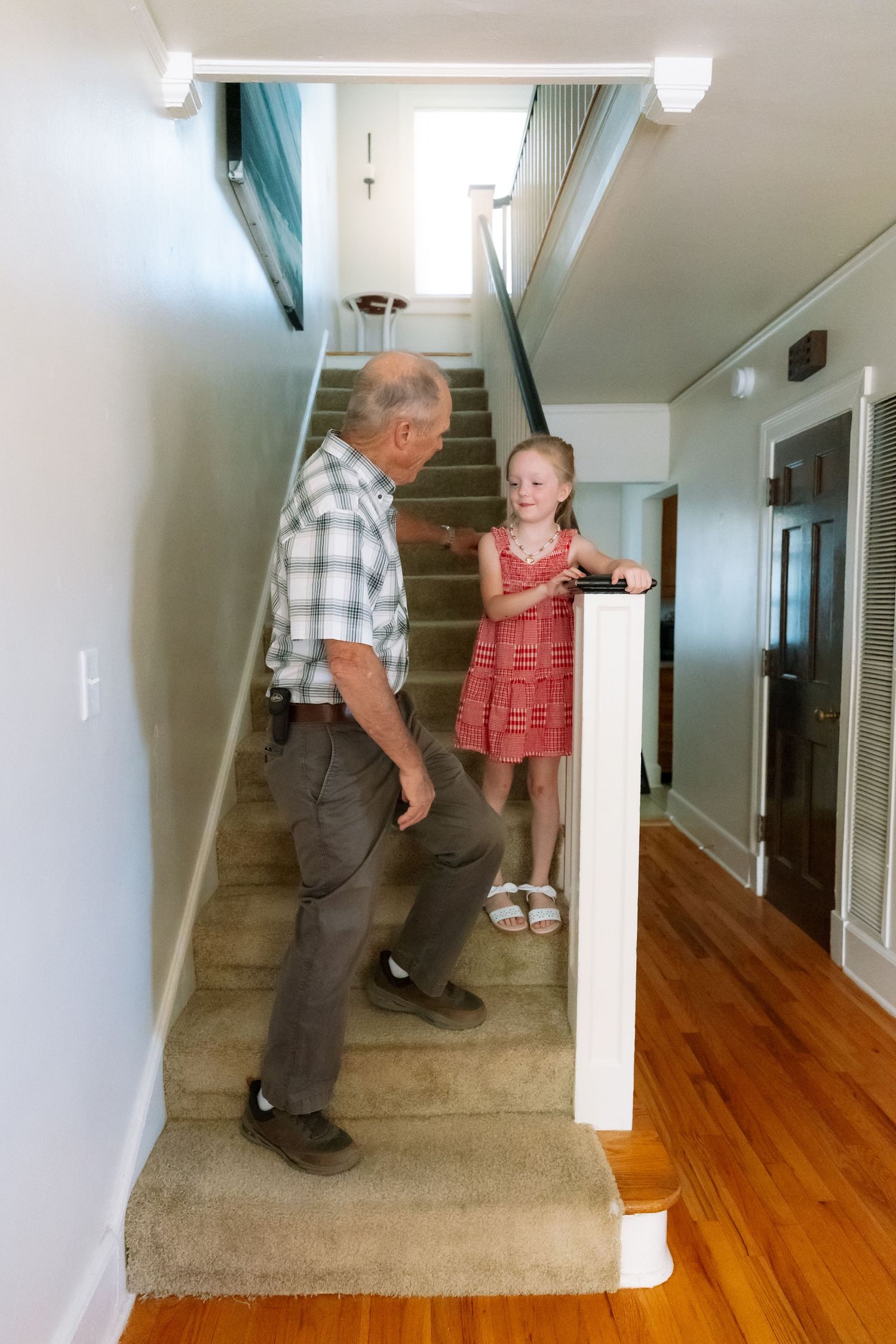 Elderly man and young girl in red dress talking on carpeted stairs inside a home with hardwood floors.