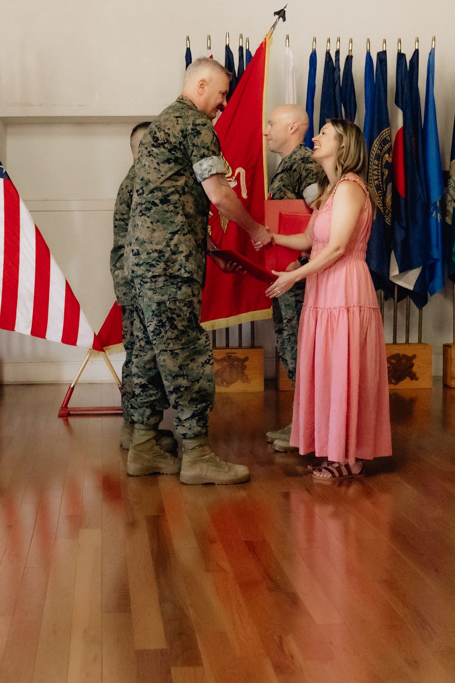 Marine officer receives a red guidon from another soldier as a woman in a pink dress assists during a military ceremony.