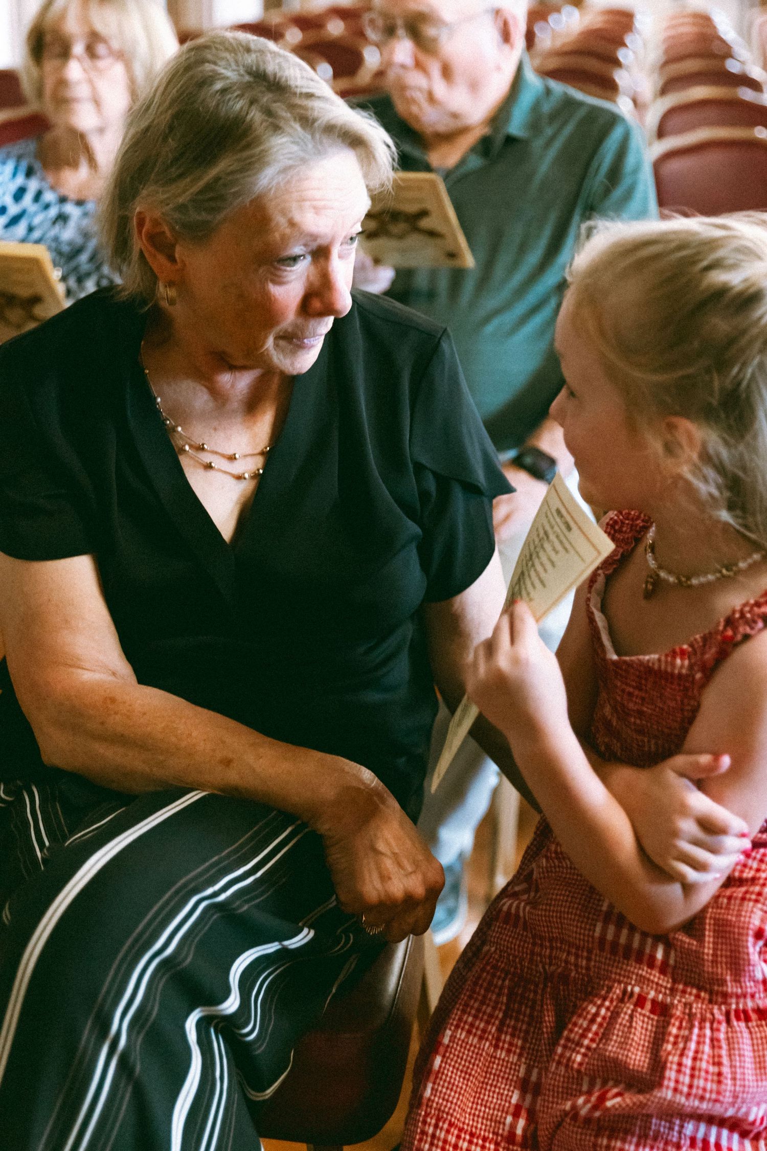 Elderly woman in black dress shares a tender moment with young girl in red dress, reading together in auditorium seating.