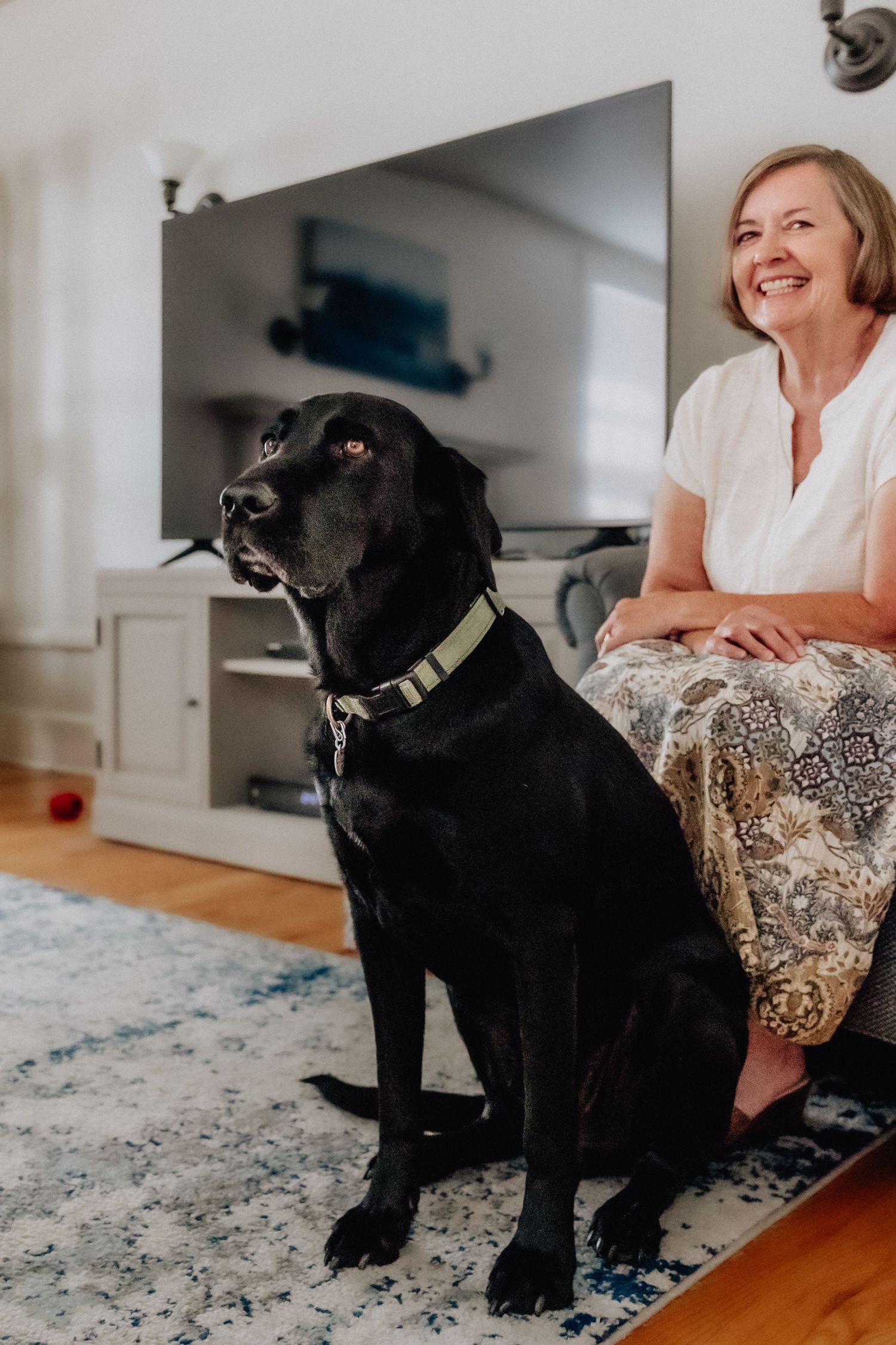 A black Labrador Retriever sits on a rug beside a smiling woman in a bright living room.