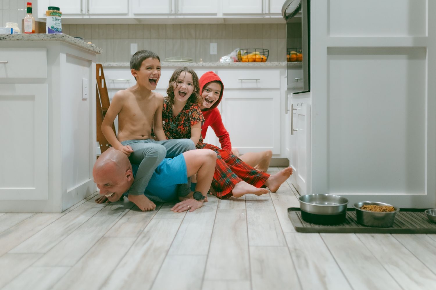 Three children laughing and sitting together on a kitchen floor, two in costumes, enjoying a playful moment at home.