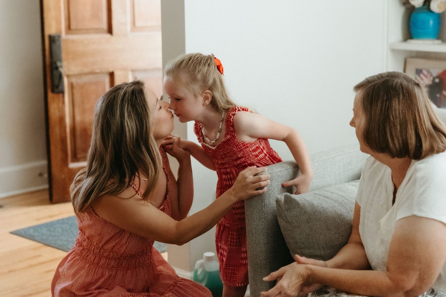 A young girl in a red dress shares a kiss with a woman while another adult watches warmly in a bright living room.